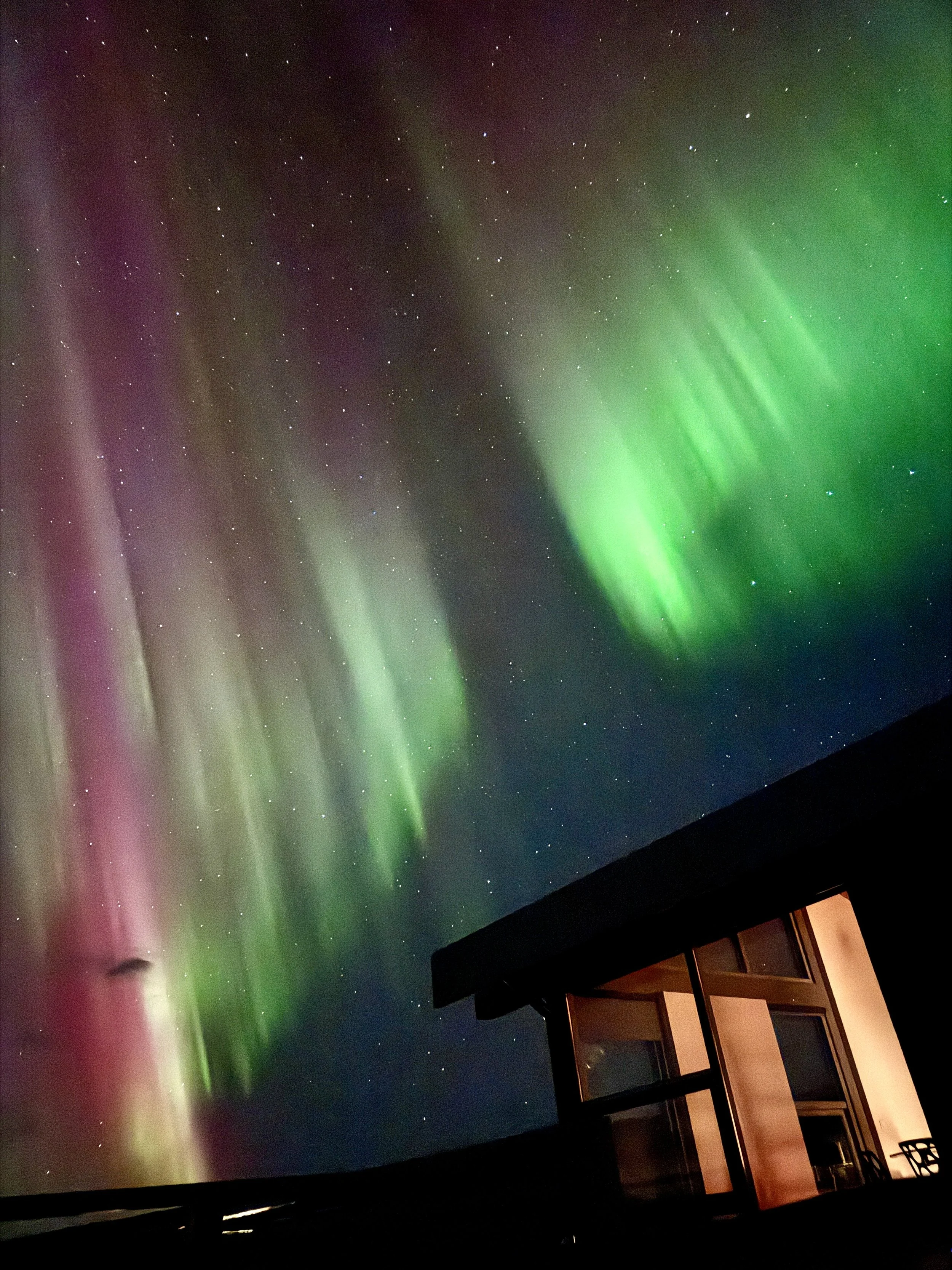 Night sky illuminated by vibrant green and purple aurora borealis over a house with a balcony.