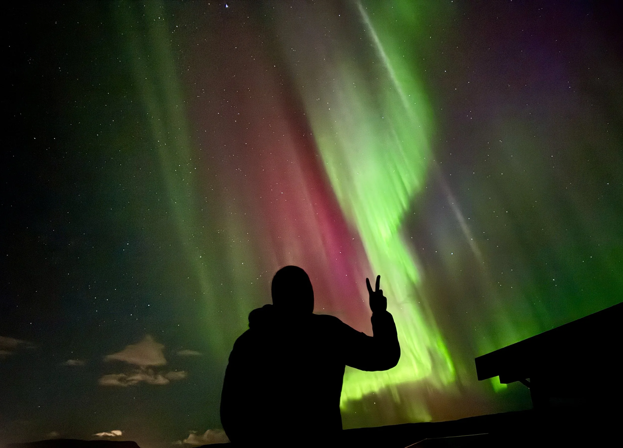 A person silhouette making a peace sign in front of the Northern Lights in the night sky.