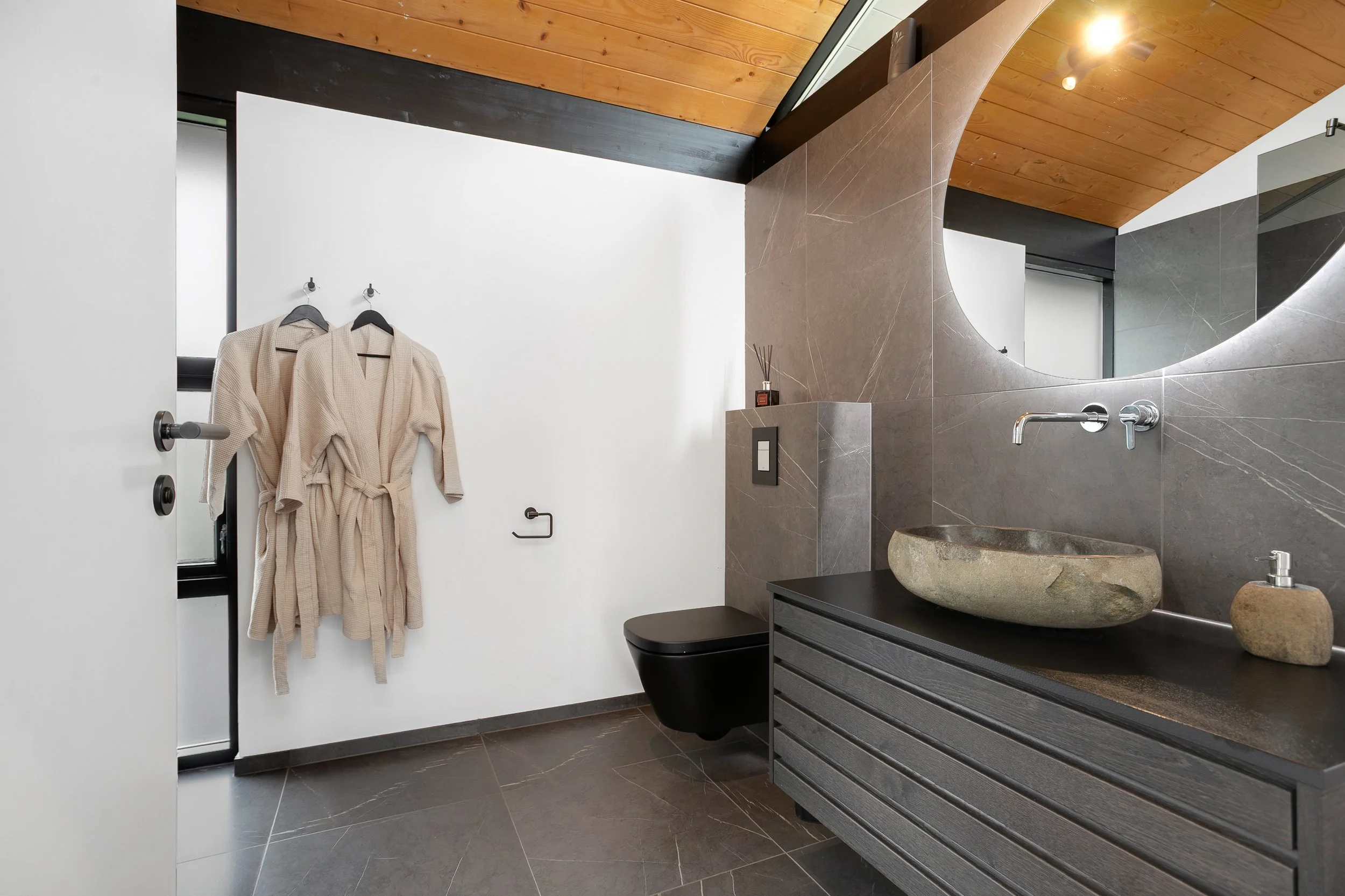 Modern bathroom featuring a wooden ceiling, a large mirror, and a stone sink on a black vanity. Beige bathrobes hang on hooks by a window, and a black toilet is visible beneath the mirror.