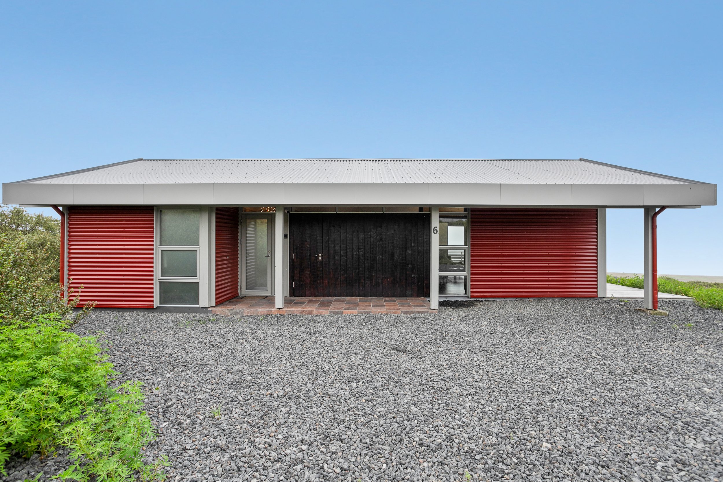 Modern house with red corrugated metal exterior, black door, grey metal roof, glass windows, gray gravel driveway, green bushes on left, and a clear blue sky.