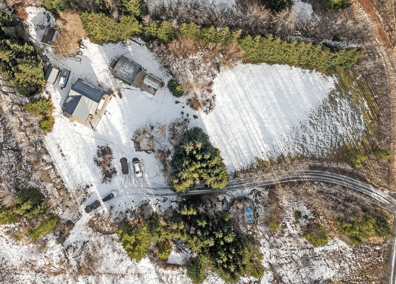 Aerial view of a snowy rural property with a house, several smaller structures, a driveway with parked cars, trees, and a large snow-covered field.