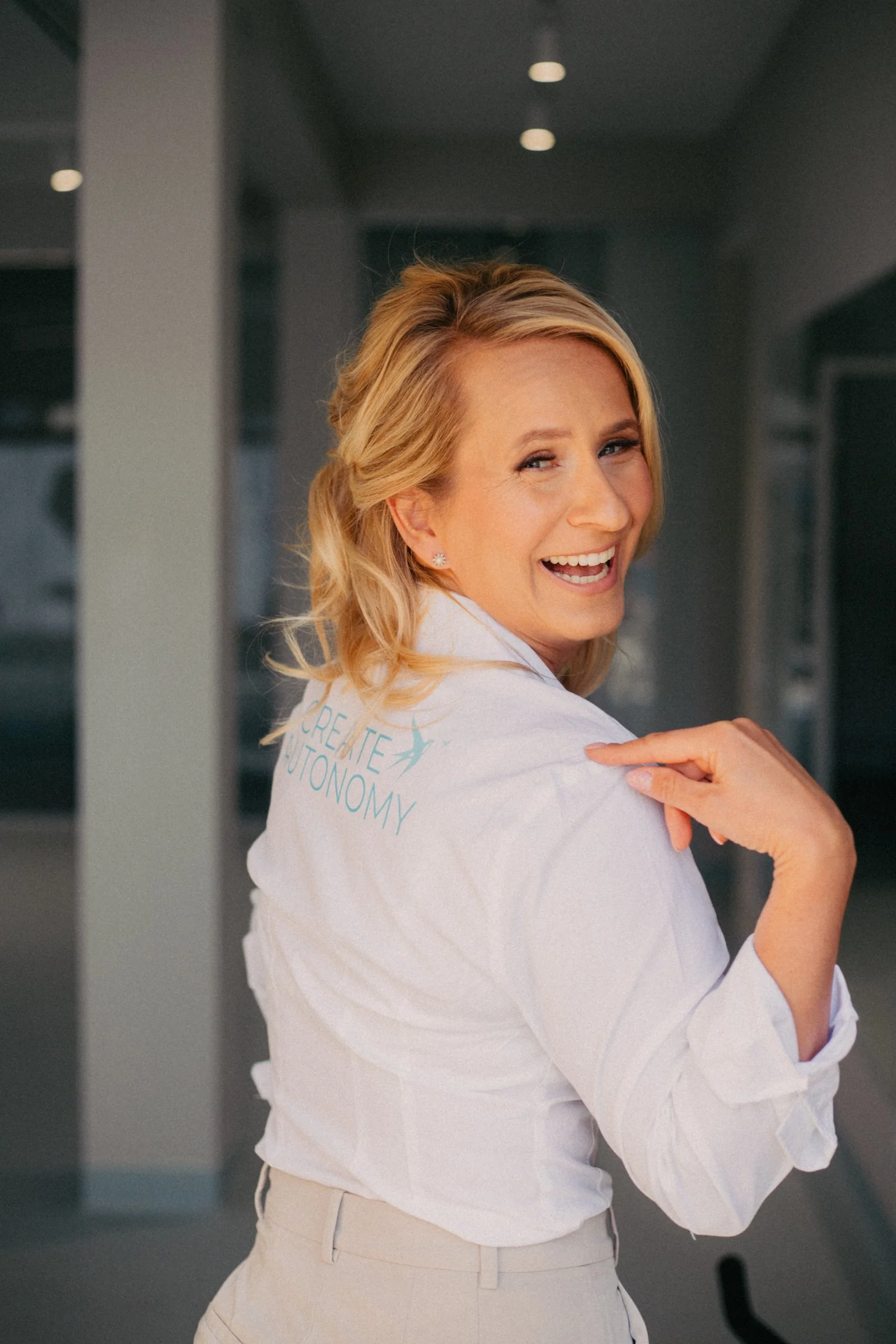 A blonde woman wearing a white shirt smiling and looking over her shoulder in a modern indoor setting.