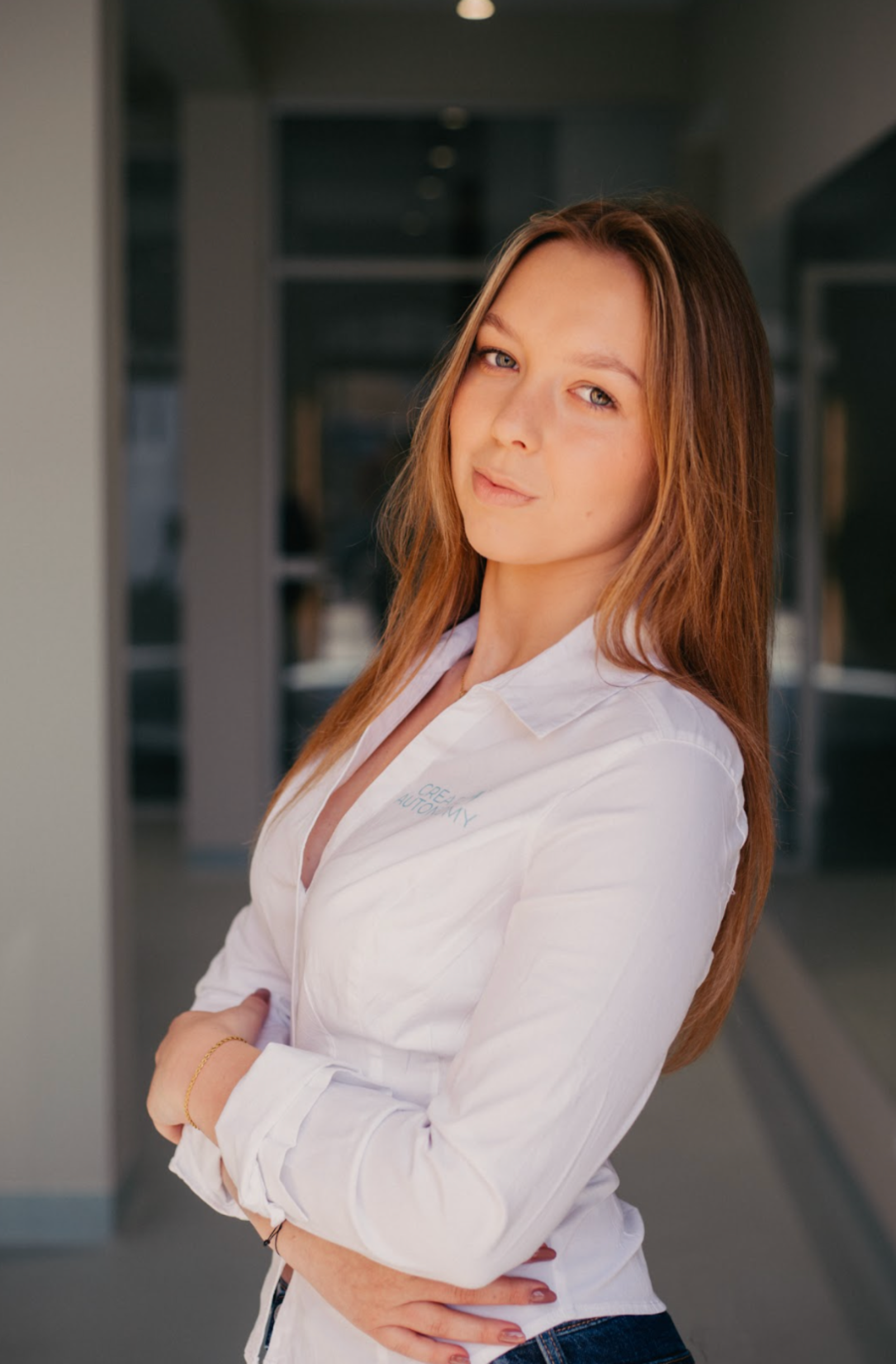A woman with long, reddish-brown hair and blue eyes standing indoors with her arms crossed, wearing a white shirt with a logo on the chest and dark jeans, in a modern building or office environment.