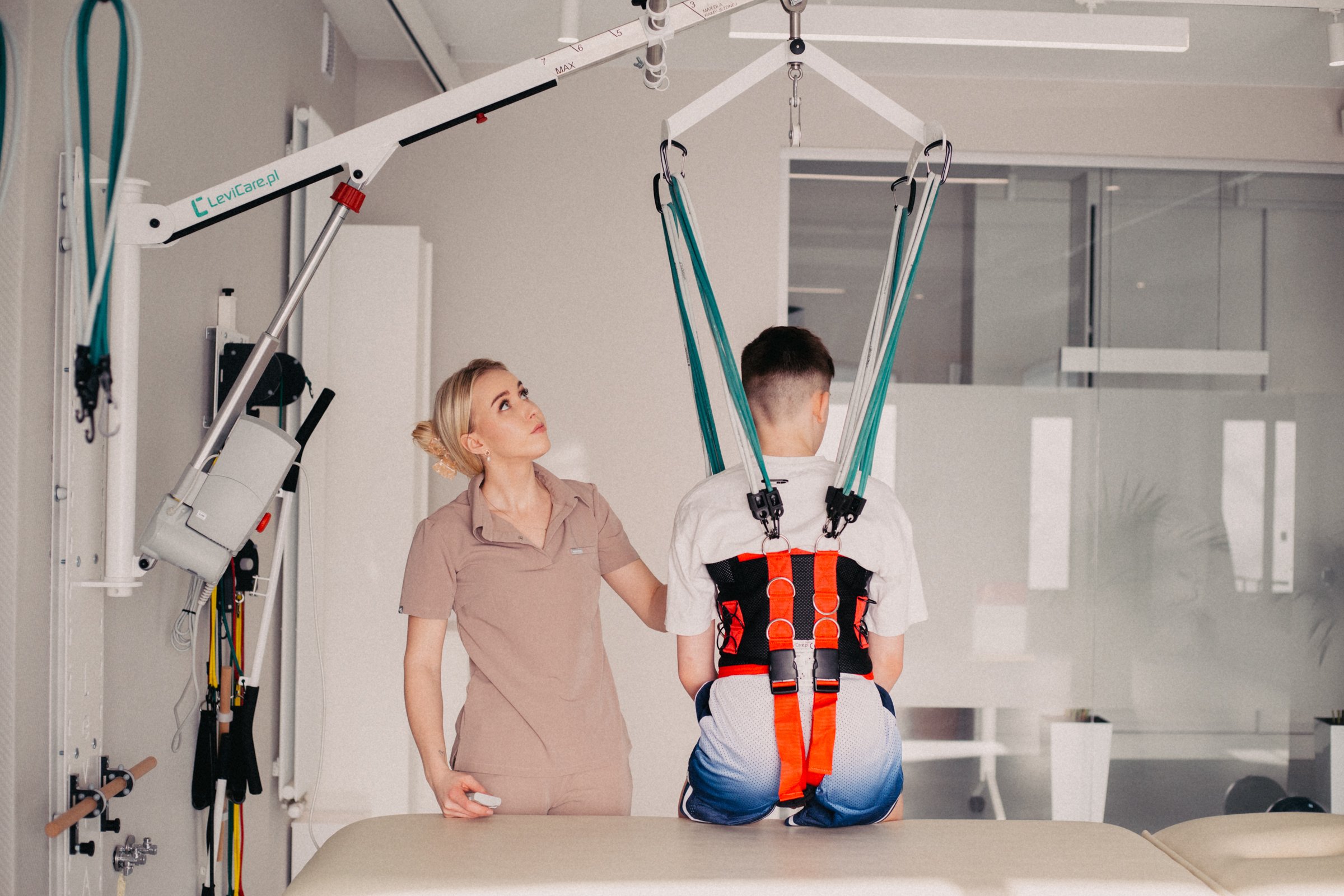 A woman assisting a man with physical therapy using a harness and pulley system, with the woman observing and standing beside him in a clinical setting.