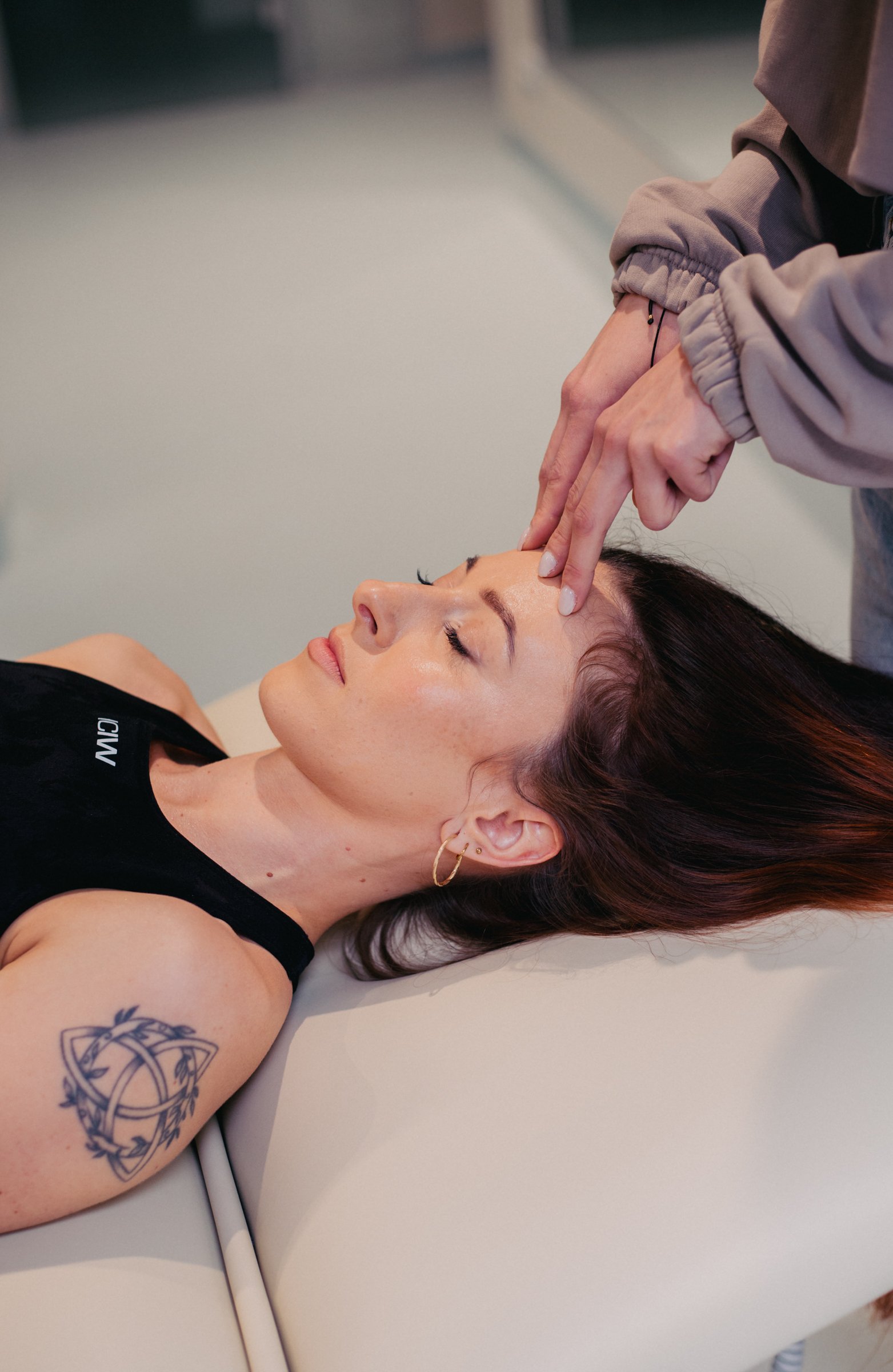 A woman with long dark hair and tattoos relaxing with her eyes closed, lying on a table while a person in a beige hoodie performs a skincare treatment on her forehead.