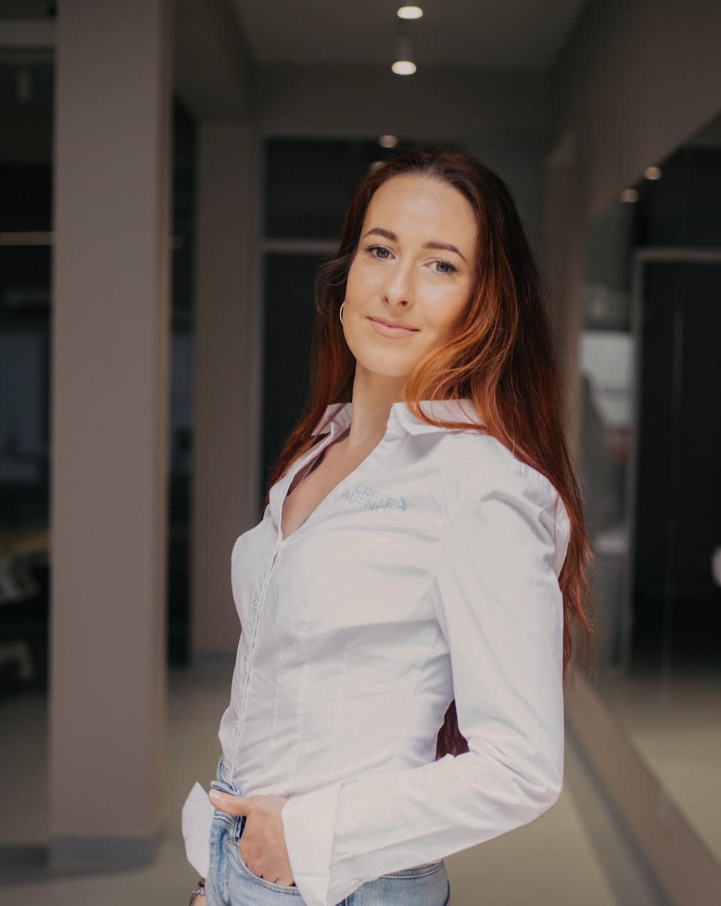 A woman with long red hair, wearing a white blouse and light blue jeans, standing indoors in a modern building with a confident expression.