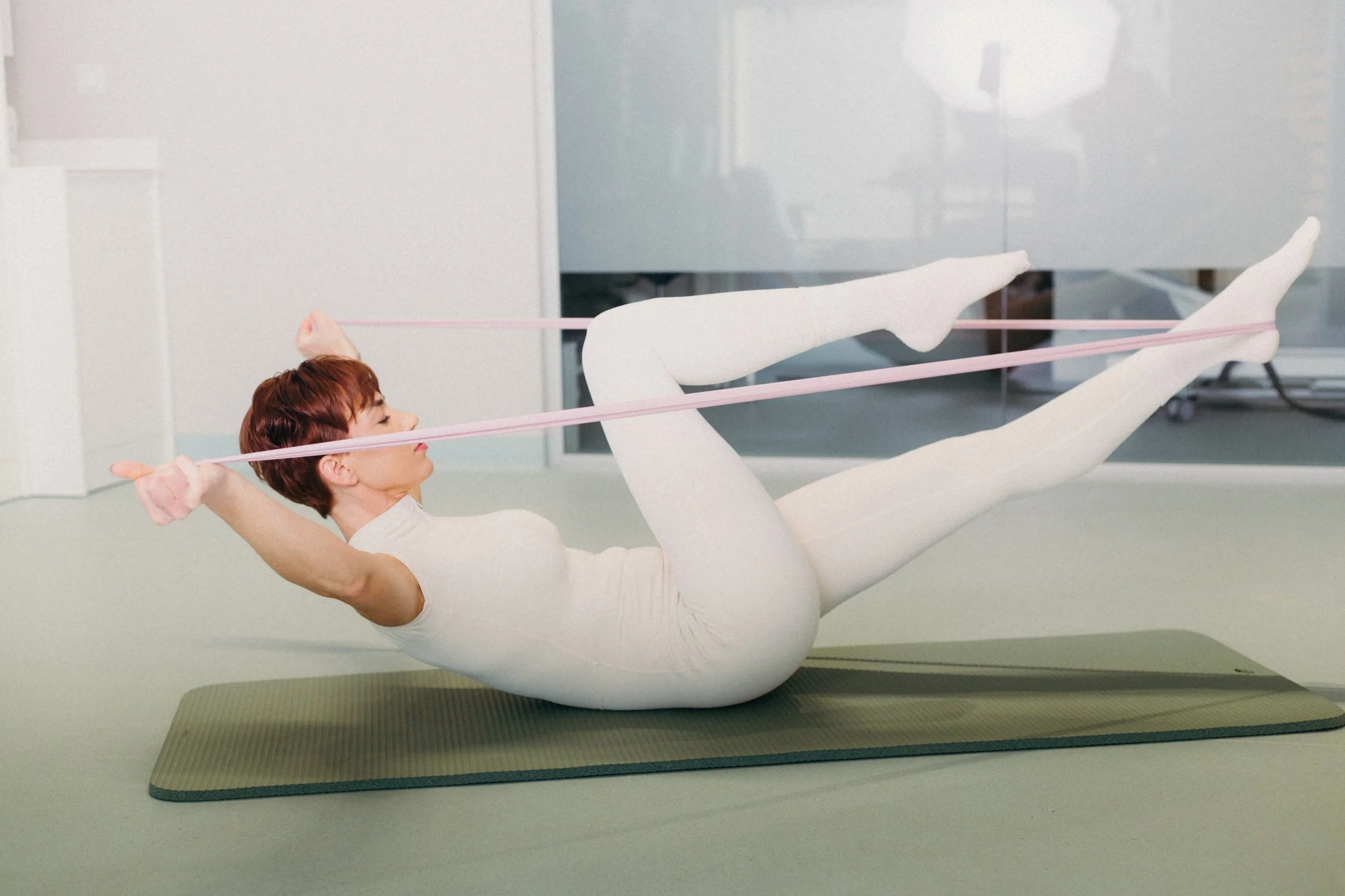 A woman with short red hair and wearing a white workout outfit is lying on a yoga mat and using a pink resistance band to stretch her legs.