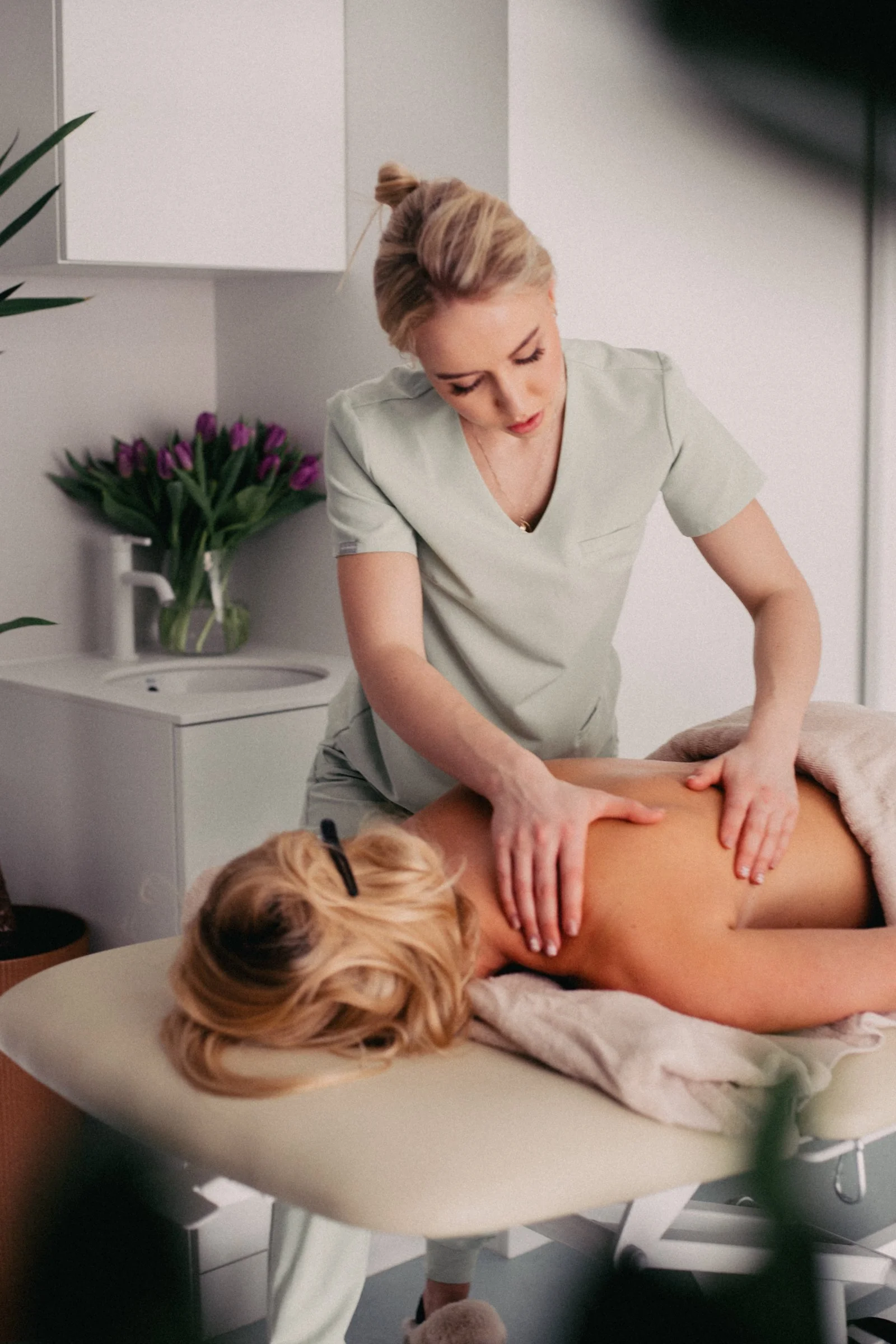 A woman receiving a massage from a therapist in a spa or massage room, with a vase of purple tulips and a white cabinet in the background.
