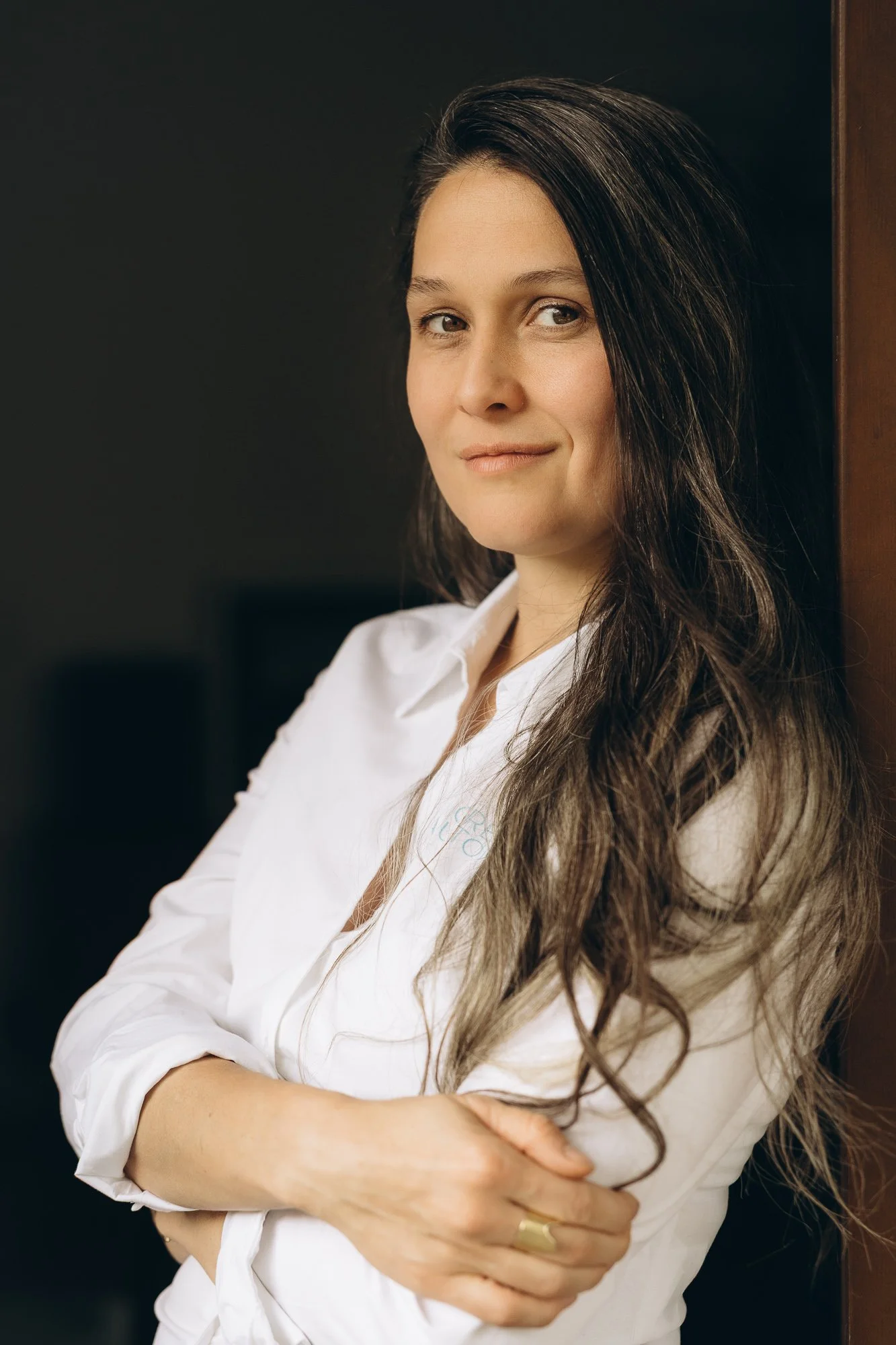 A woman with long, wavy brown hair wearing a white shirt is standing against a dark background, looking at the camera with a slight smile and crossed arms.