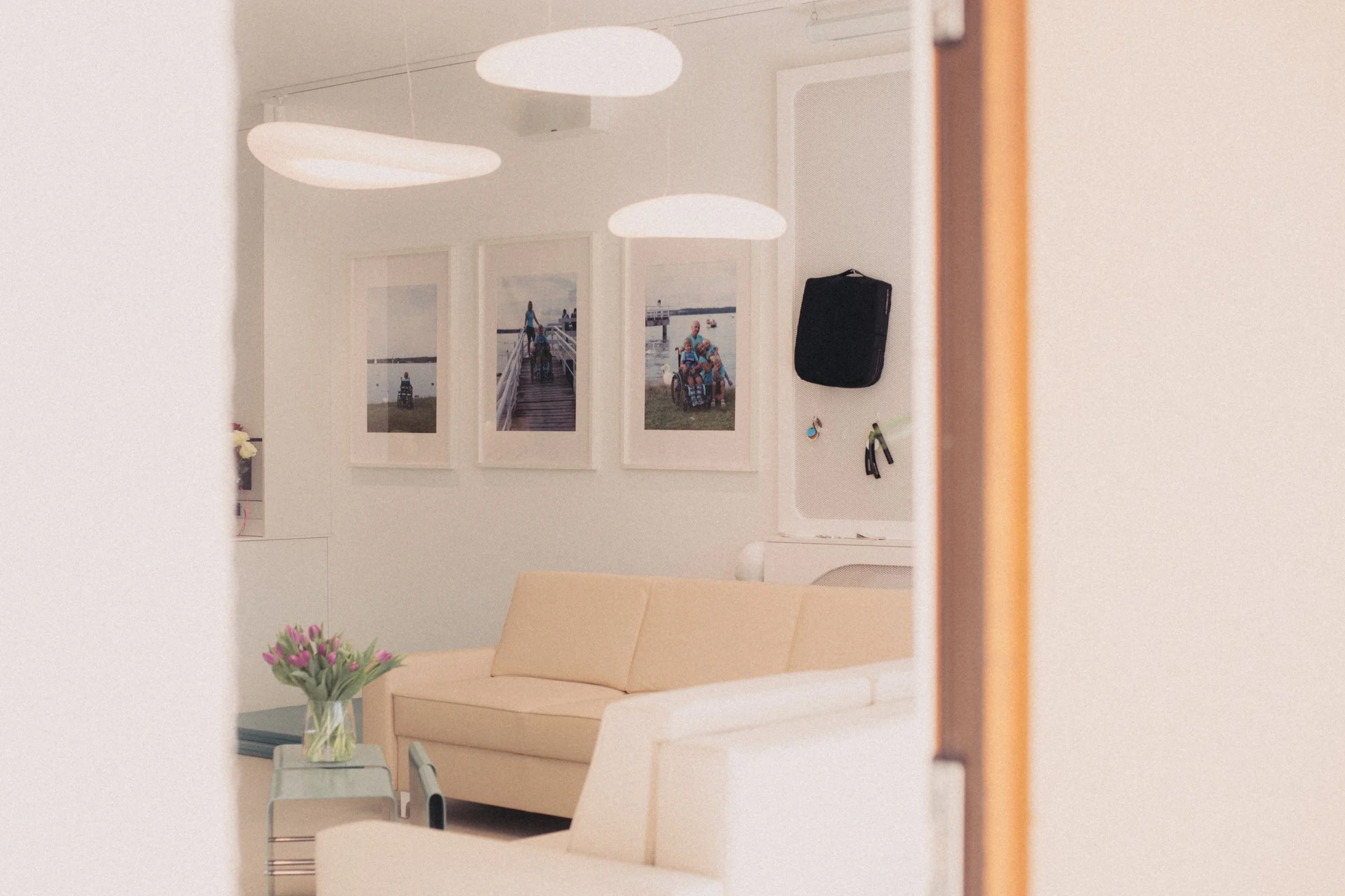 A reflection of a room with a beige sofa, a glass vase with pink flowers, framed pictures on the wall, and a black bag hanging on the wall in a mirror.
