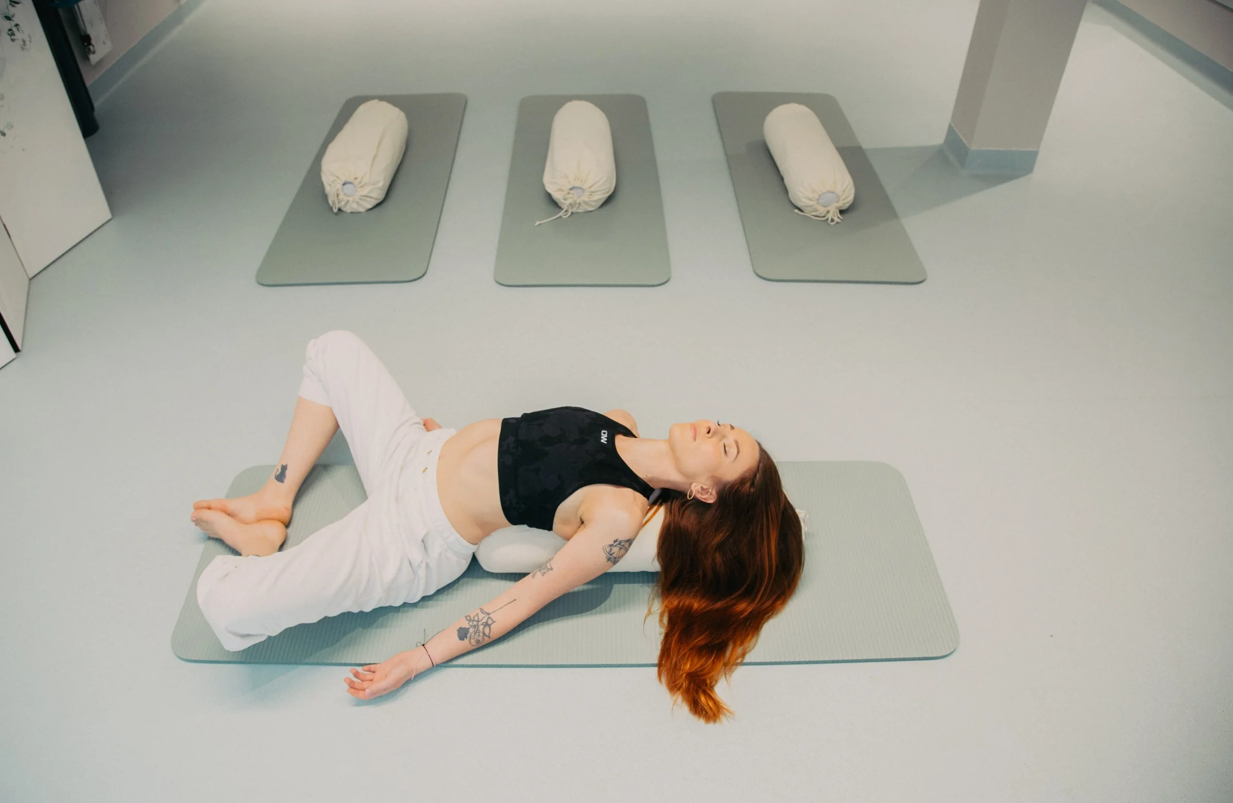 A woman with red hair lying on a yoga mat in a hospital room, appearing to be practicing relaxation or meditation. There are three other yoga mats with rolled-up mats on top of them in the background.