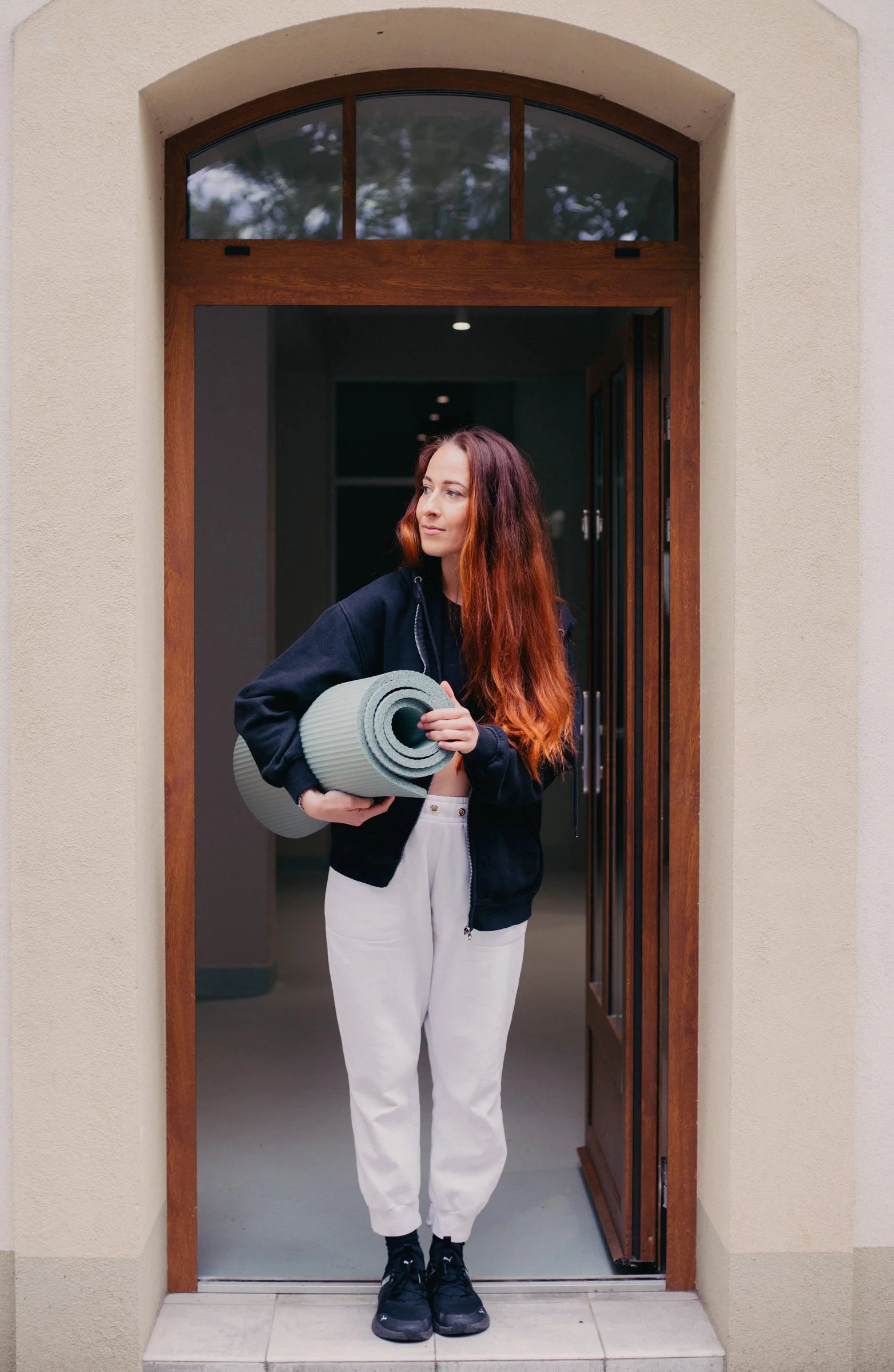 A young woman with long reddish hair, dressed in white pants and a dark jacket, holding a rolled-up yoga mat, standing in a doorway of a house.