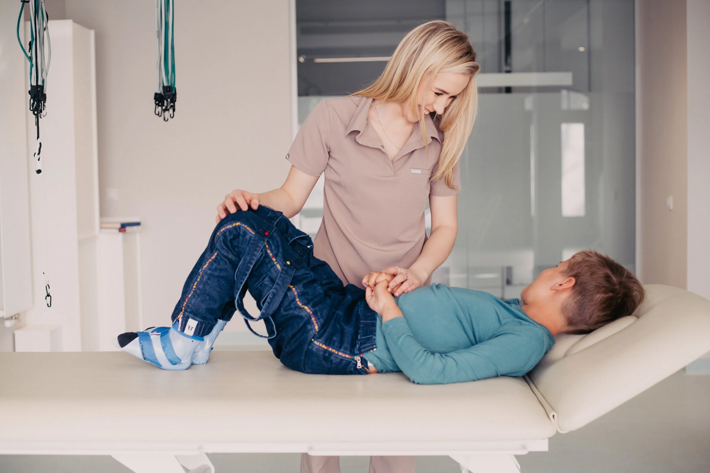 A young female nurse smiling and helping a boy who is lying on an examination bed in a hospital or clinic setting.