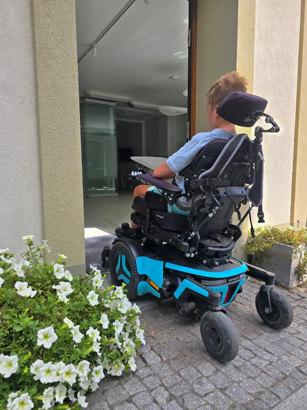 A young boy in a motorized wheelchair is outside on a paved area, looking towards a building entrance with large glass doors. There are white flowers in a planter to the left.
