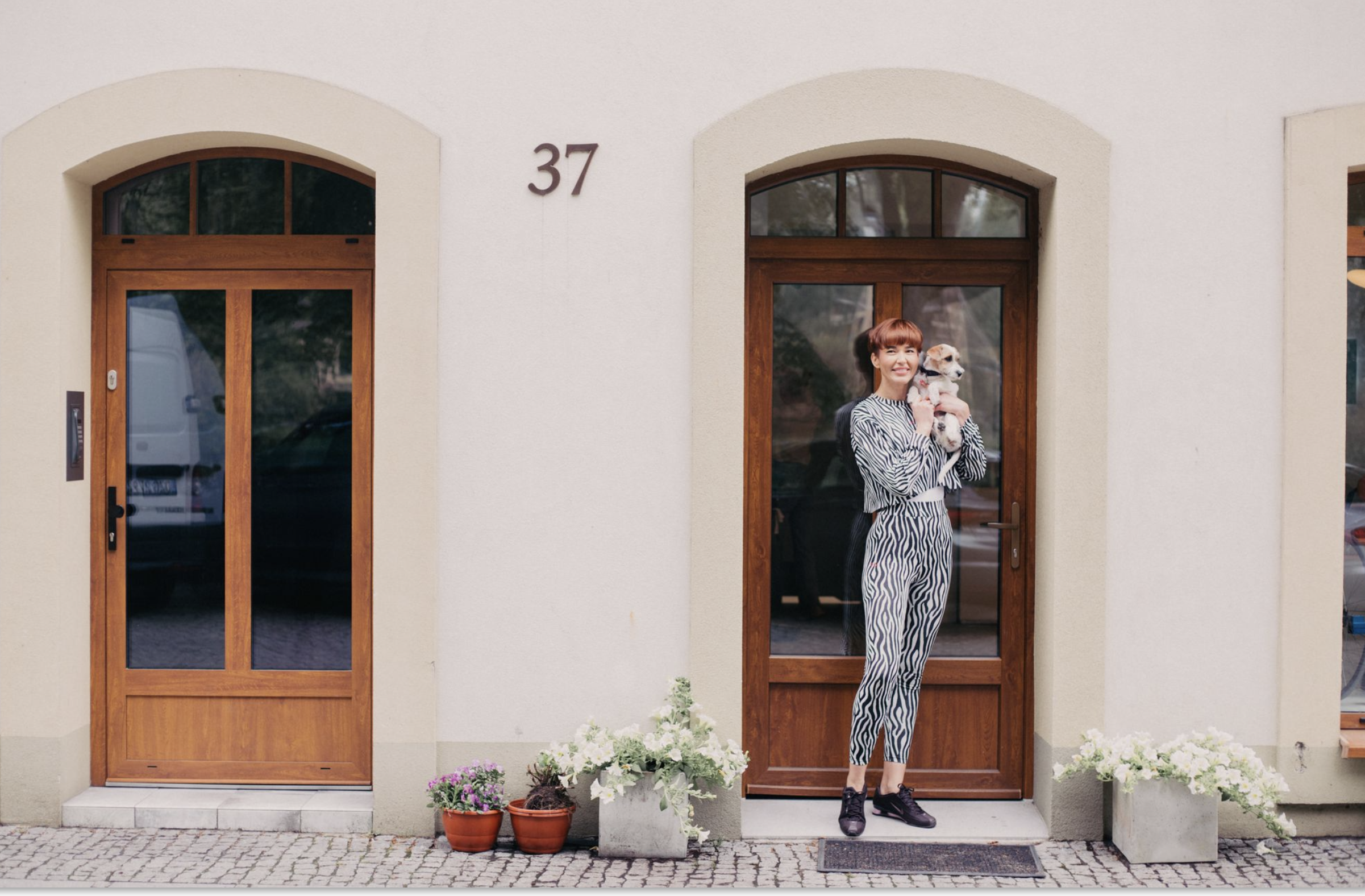 A woman with short hair wearing a zebra print dress and sneakers holding a small dog, standing in front of a house with a beige facade and wooden framed glass doors, with house number 37 and potted plants with white and purple flowers.