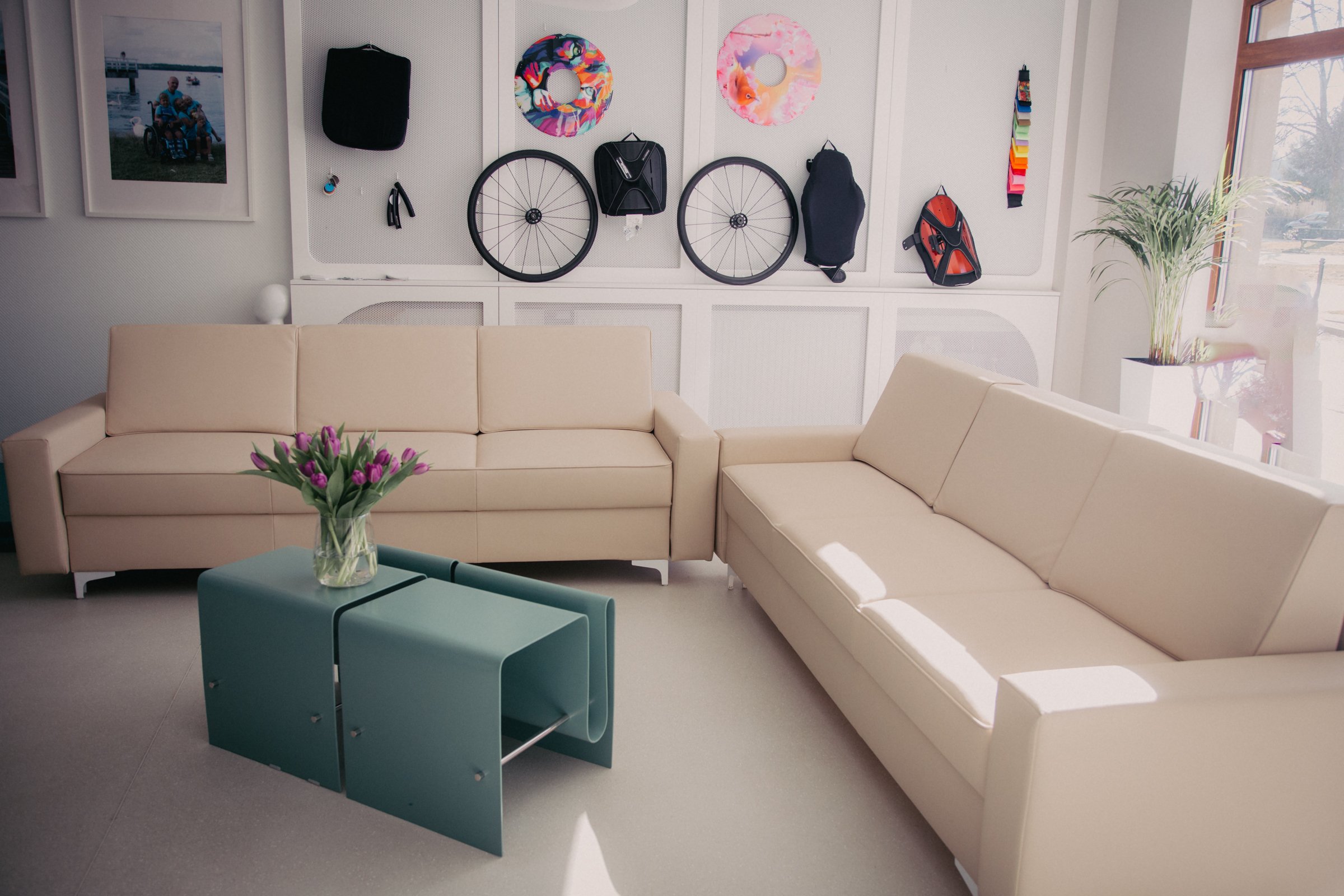A cozy waiting room with a beige sectional sofa, a coffee table with a vase of purple tulips, and a wall displaying bicycle wheels, colorful floats, and a photograph of a family at the beach. A tall plant is near a window on the right.