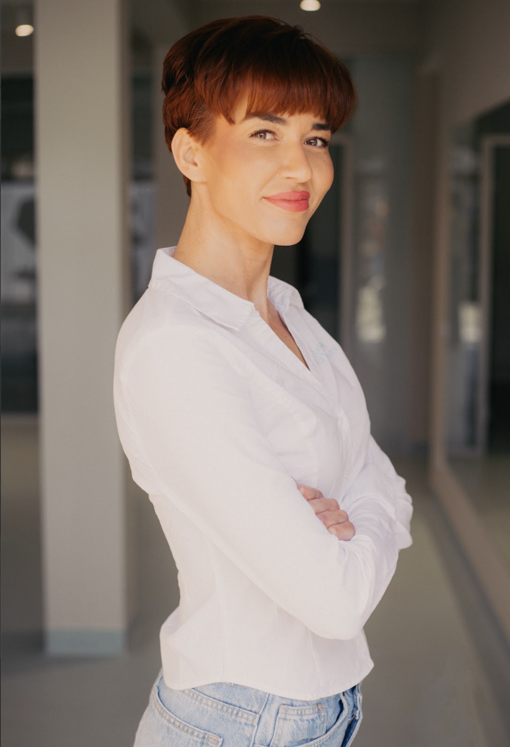 A woman with short brown hair and a white shirt standing with her arms crossed in an indoor setting.
