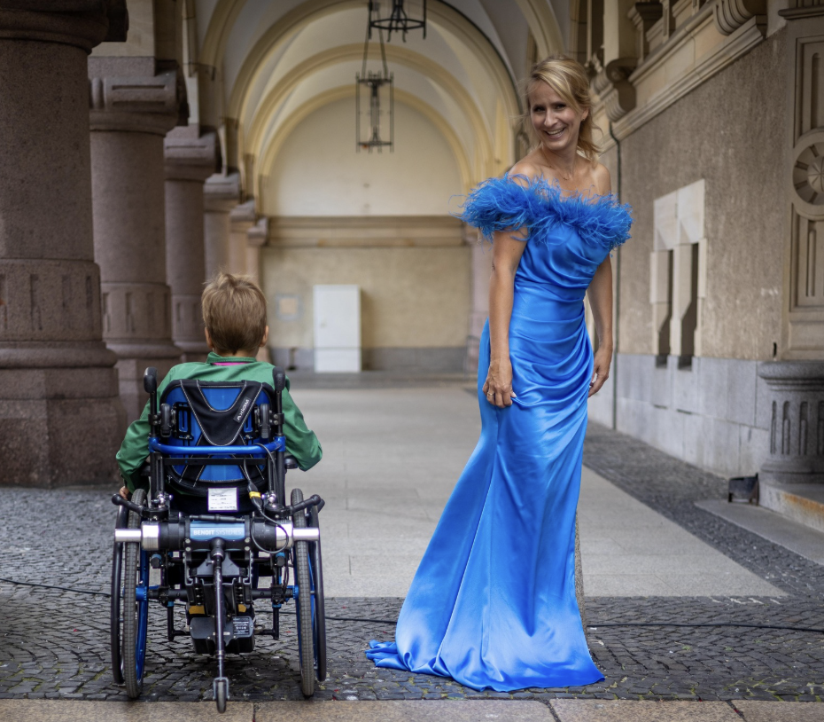 A woman in a bright blue, off-the-shoulder gown with feather embellishments standing and smiling near a young boy in a wheelchair in a covered outdoor area with stone architecture.
