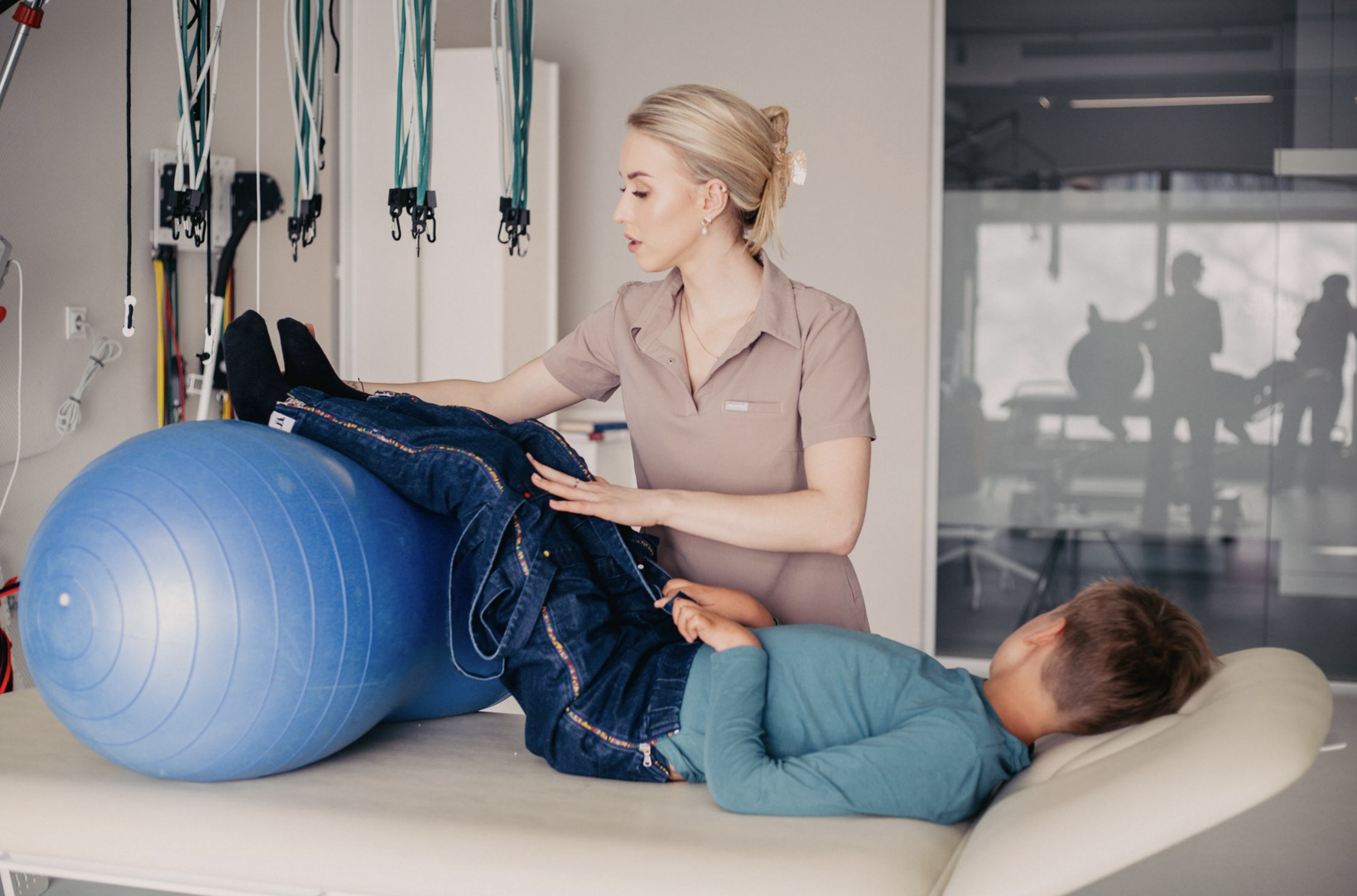 A woman in a beige uniform assisting children with physical therapy exercises on an exercise ball in a rehabilitation center.