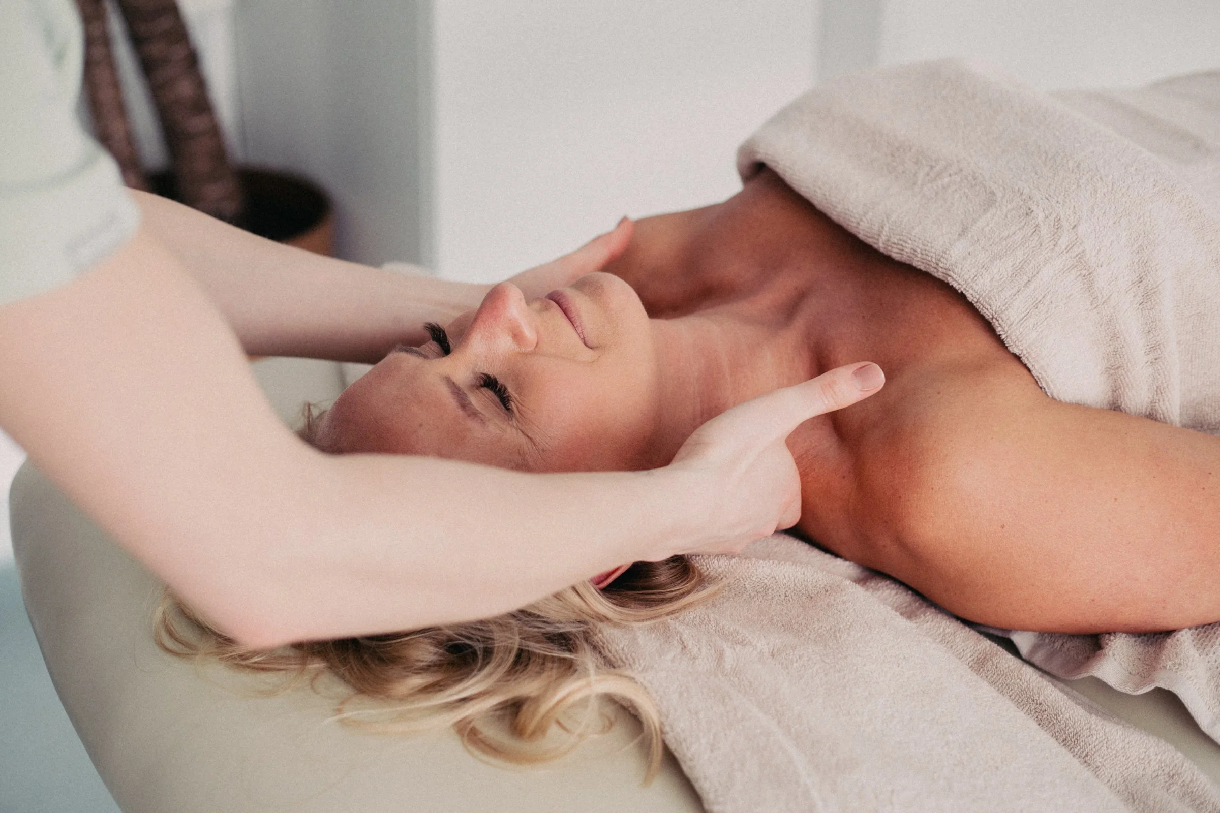 A woman lying on a massage table with her eyes closed, receiving a massage therapy from a therapist. The woman has blonde hair and appears relaxed.
