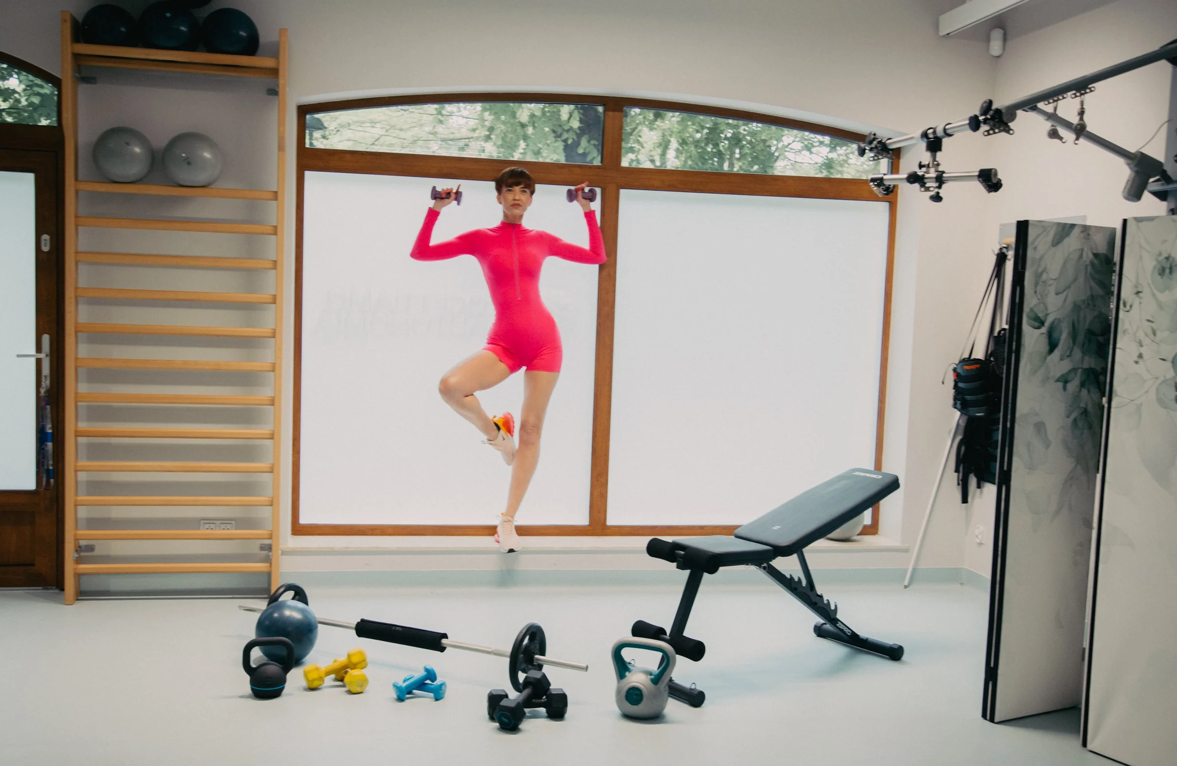 Woman in red workout suit exercising with dumbbells in a home gym with various exercise equipment and large windows.