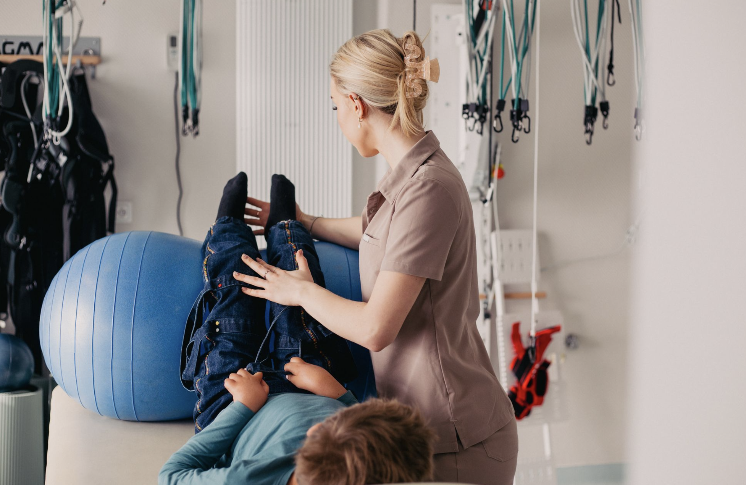A woman in a beige uniform assisting a young boy with a physical therapy exercise using a large blue exercise ball in a healthcare or therapy setting.