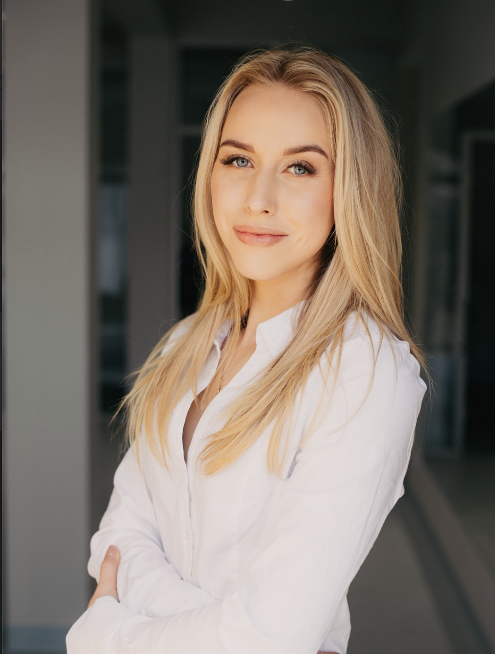 A young woman with blonde hair and blue eyes smiling softly, wearing a white shirt, standing outside in daylight.