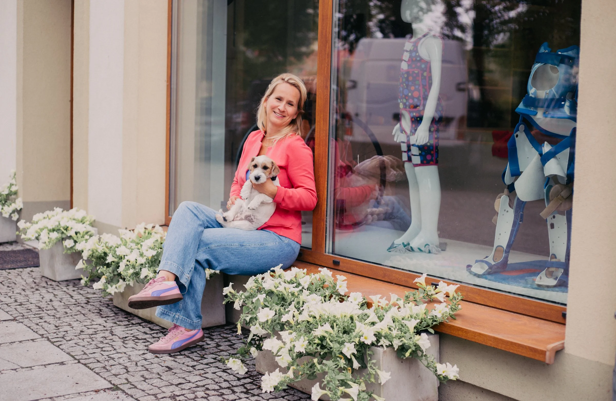 A woman sitting outside a shop window holding a small puppy and smiling. The shop window displays a mannequin wearing a colorful polka-dotted dress and a pair of robotic legs. There are white flowers in rectangular planters along the sidewalk.