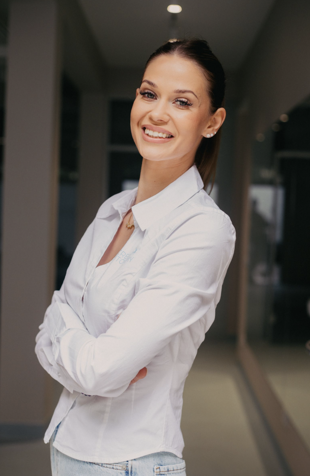 A smiling woman with dark hair in a ponytail wearing a white button-up shirt and light denim jeans standing indoors with arms crossed.