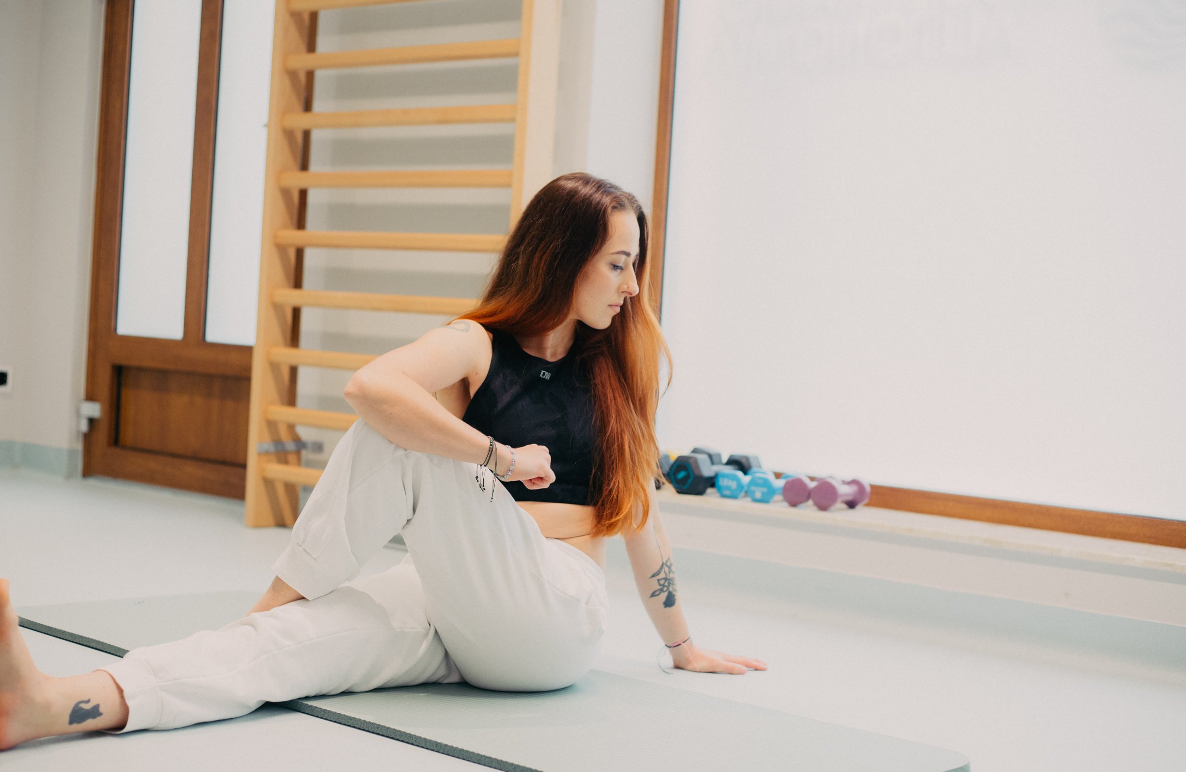 A woman with long reddish-brown hair doing yoga in a gym. She is sitting on a yoga mat, wearing a black sports bra and white pants. In the background, there are dumbbells and a wooden wall bar.