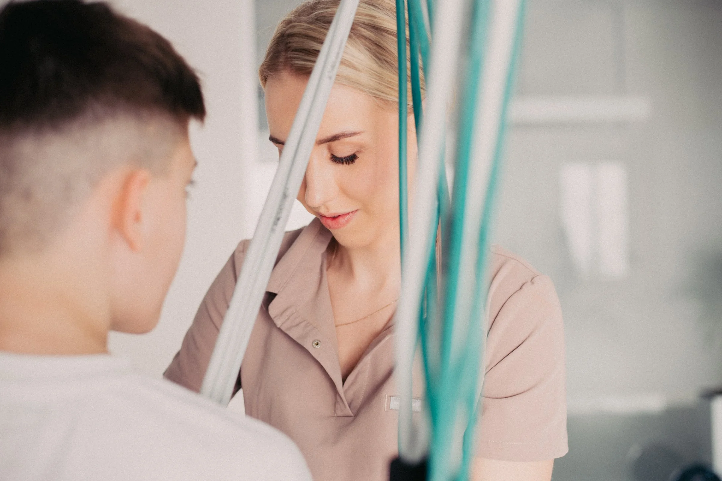 A female healthcare worker with blonde hair and a beige uniform is looking downward, with her eyes closed. In the foreground, a young man with short dark hair, seen from behind, appears to be a patient. The setting looks like a medical or clinical environment, with IV or other medical tubes visible.