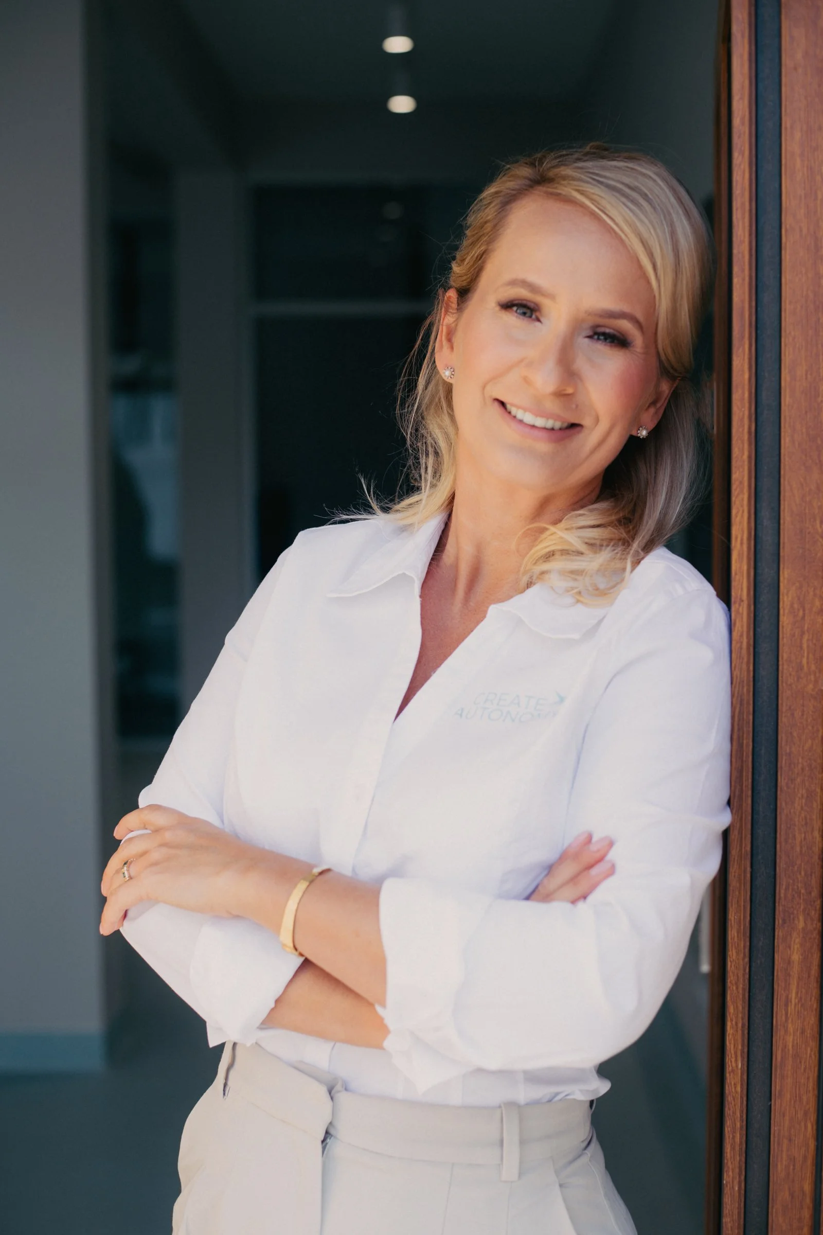 A woman with blonde hair smiling and crossing her arms, wearing a white shirt and light-colored pants, standing next to a wooden wall.