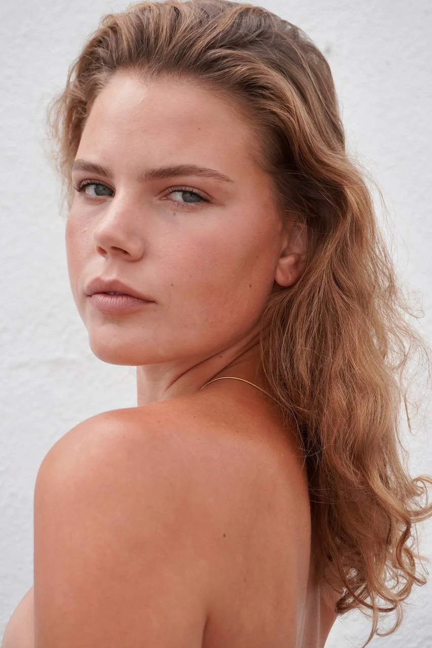 Close-up of a young woman with reddish-brown, wavy hair and blue eyes against a light background.
