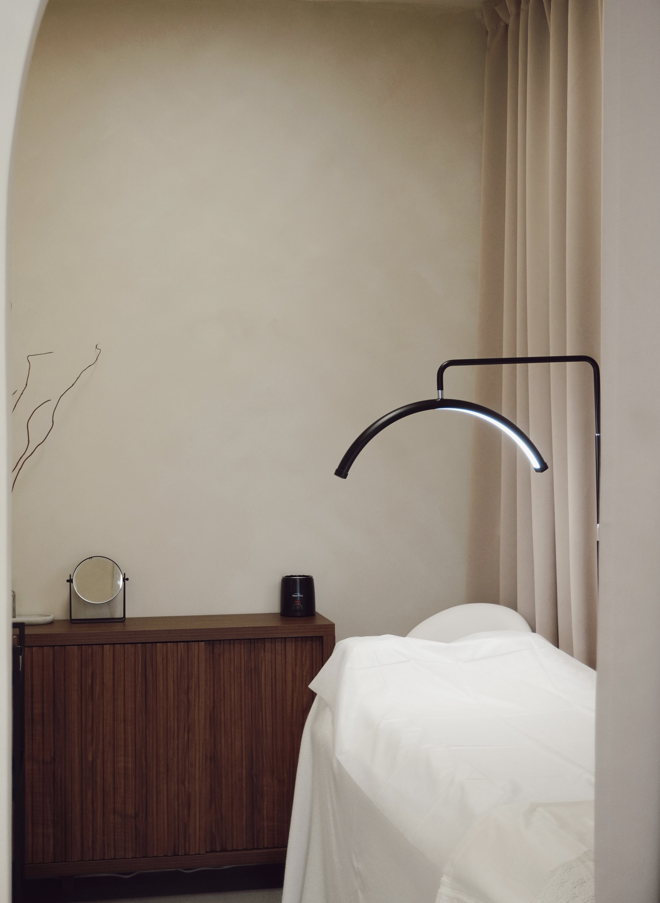 A bedroom with a bed partially covered in white sheets, a wooden nightstand with a small mirror, a black speaker, and a small black bottle. A black arc-shaped lamp hangs above the bed. Cream-colored curtains are on the right side of the room, and a cream-colored wall is in the background.