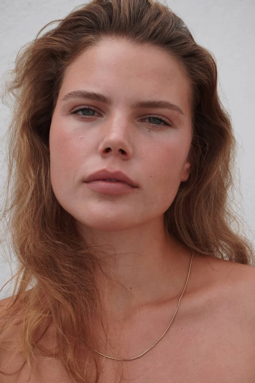 A close-up portrait of a woman with wavy red hair, blue eyes, and fair skin, wearing a thin gold necklace against a plain light background.
