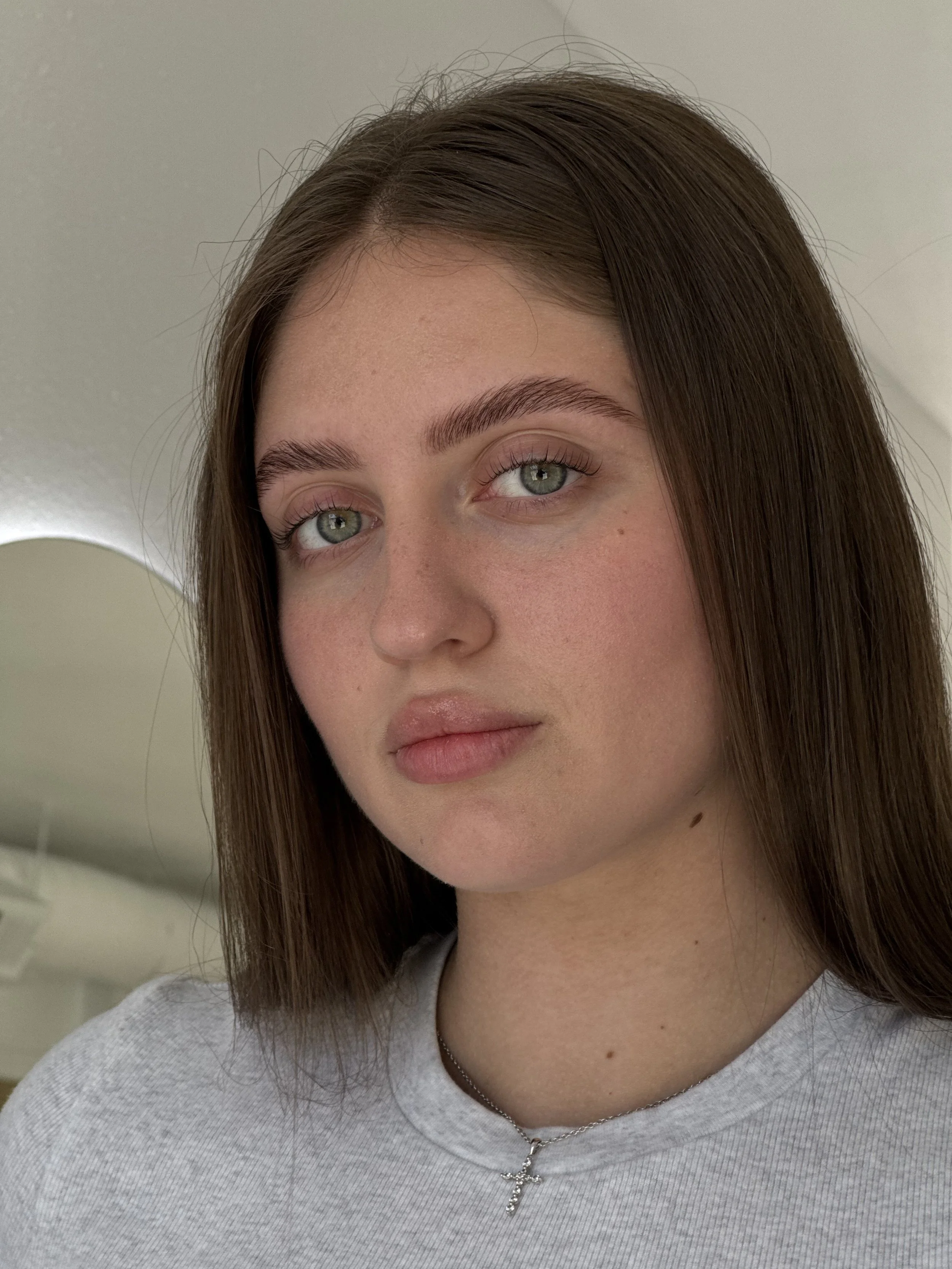 Close-up of a young woman with brown hair, green eyes, wearing a gray top, a silver cross pendant necklace, looking at the camera.