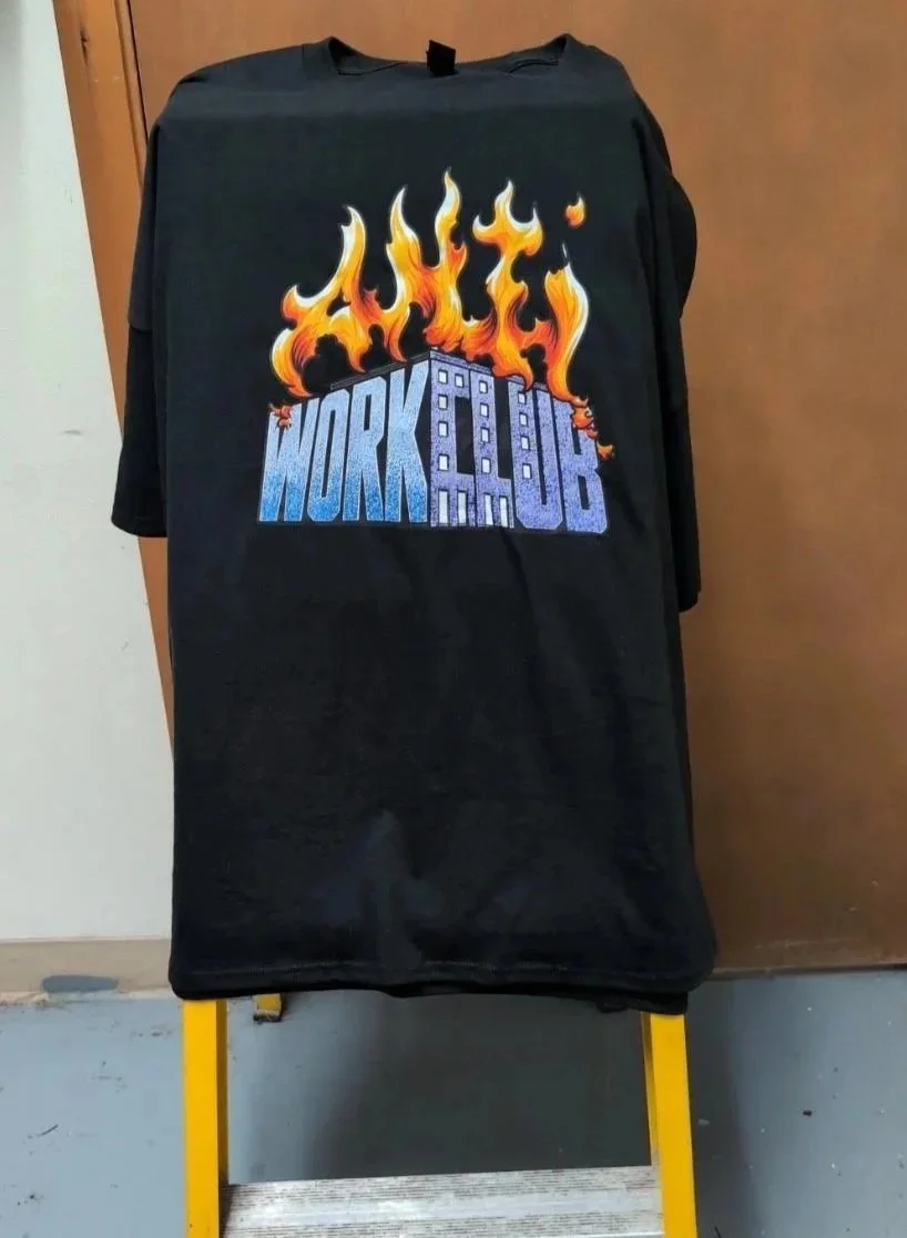 Black T-shirt with a colorful graphic design featuring flames and the words "WORK A JOB" in stylized text, displayed on a yellow stand against a brown background.