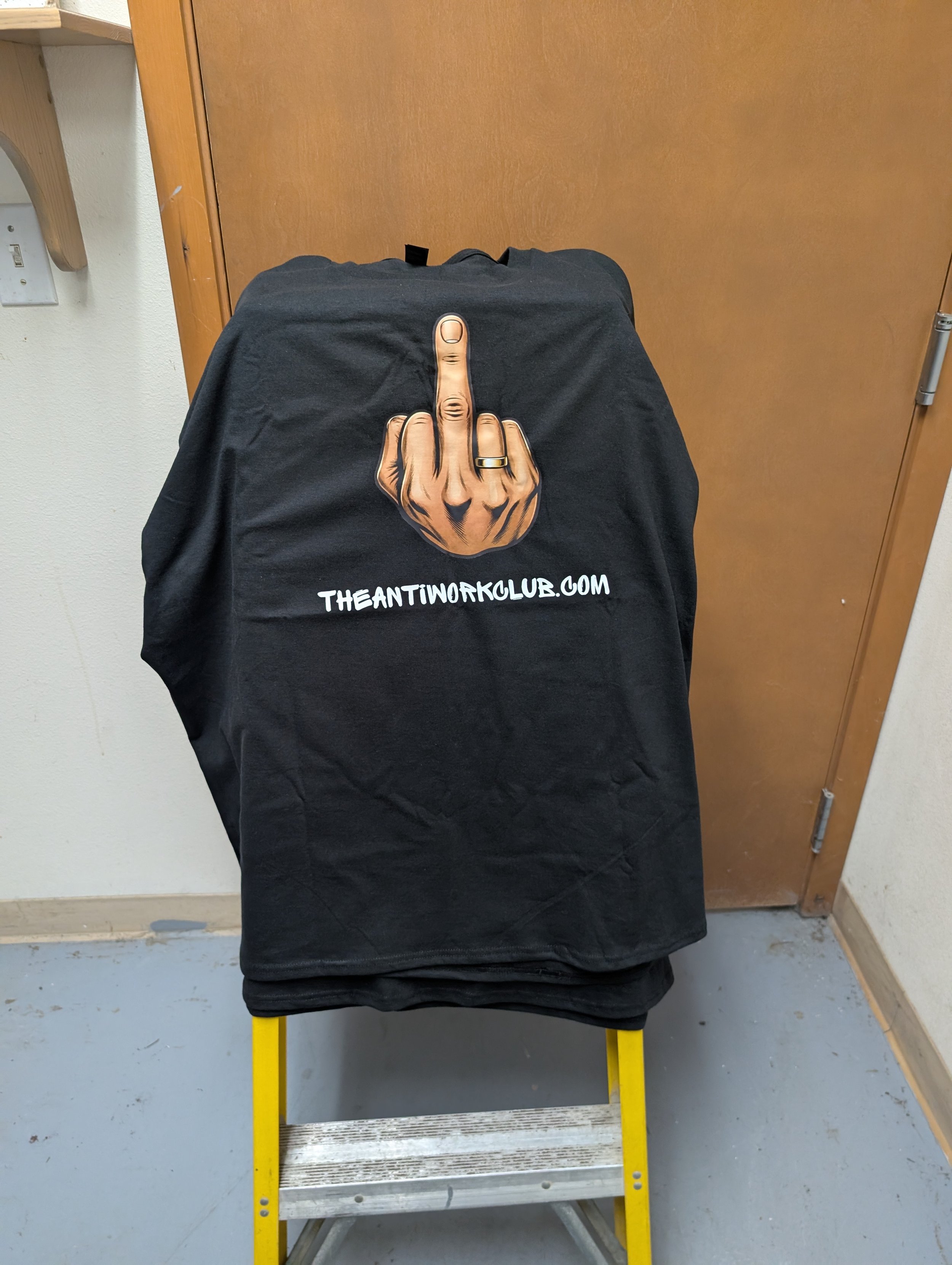 A black T-shirt hanging on a yellow step ladder. The shirt has a graphic of a hand showing the middle finger, with the text 'THEANTINORKOLUB.COM' below it. The background consists of a wooden door and a white wall.