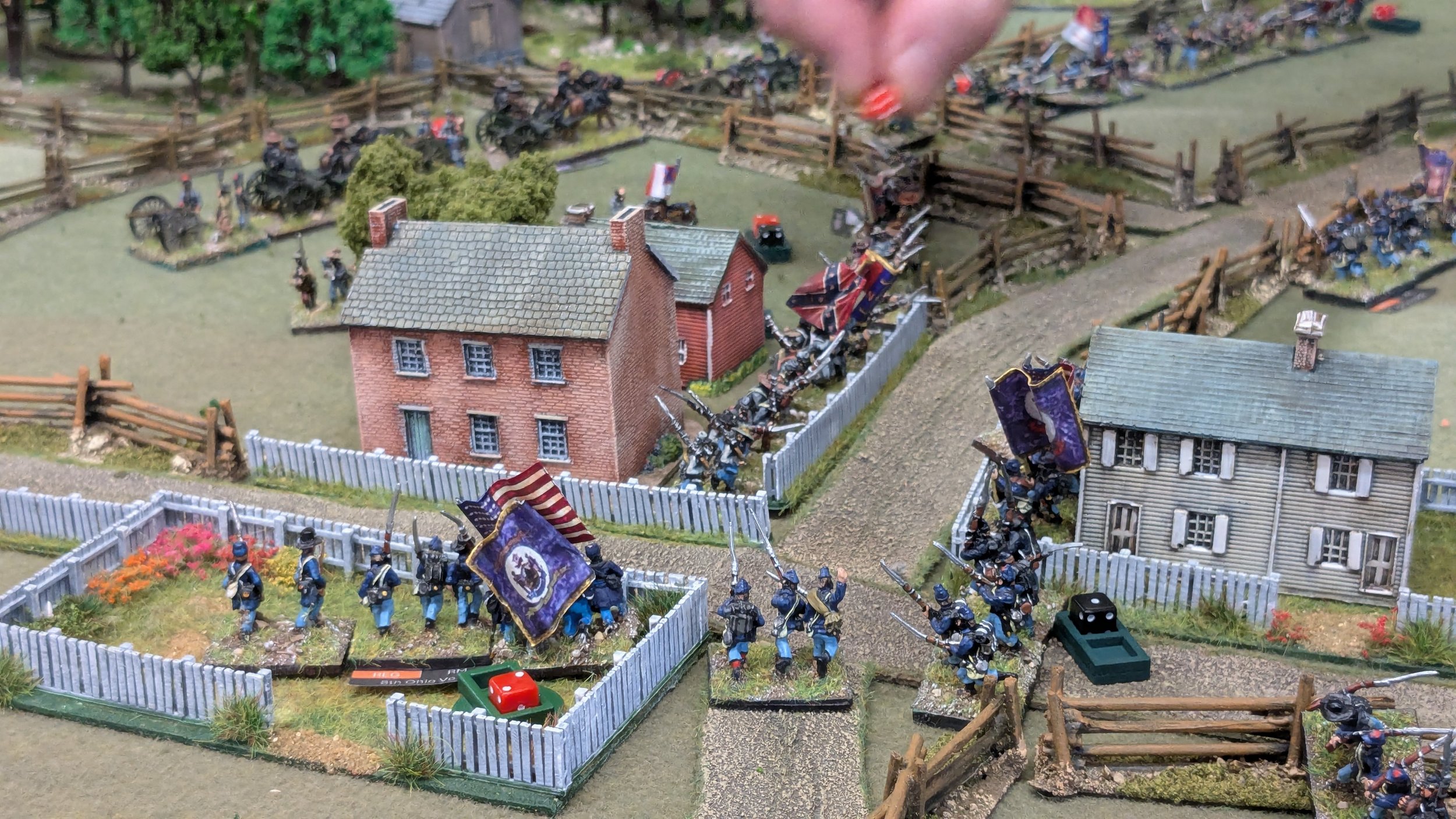 Miniature diorama depicting a historical battle scene with soldiers, cannons, and buildings, set in a rural area with fences and trees.