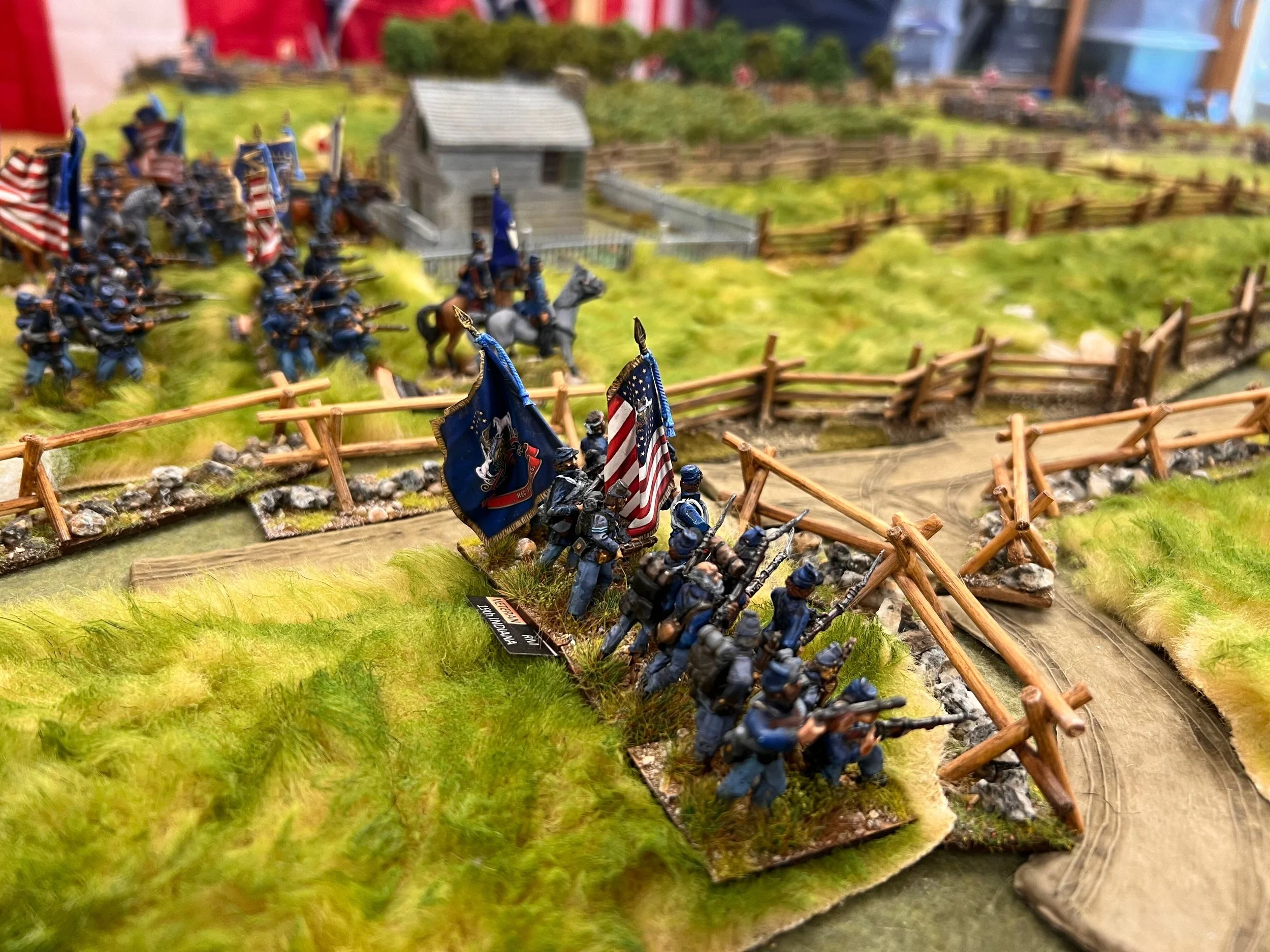 Miniature figures of soldiers in battle formation, holding flags, positioned behind a wooden fence on a grassy terrain, with a small building and additional figures in the background.