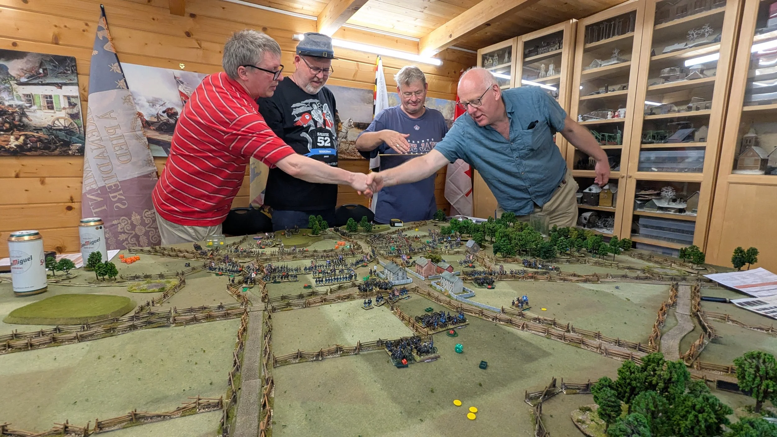 Four men are gathered around a detailed miniature battlefield in a wood-paneled room, shaking hands. The scene includes miniature trees, buildings, and soldiers, with shelves of model displays in the background.