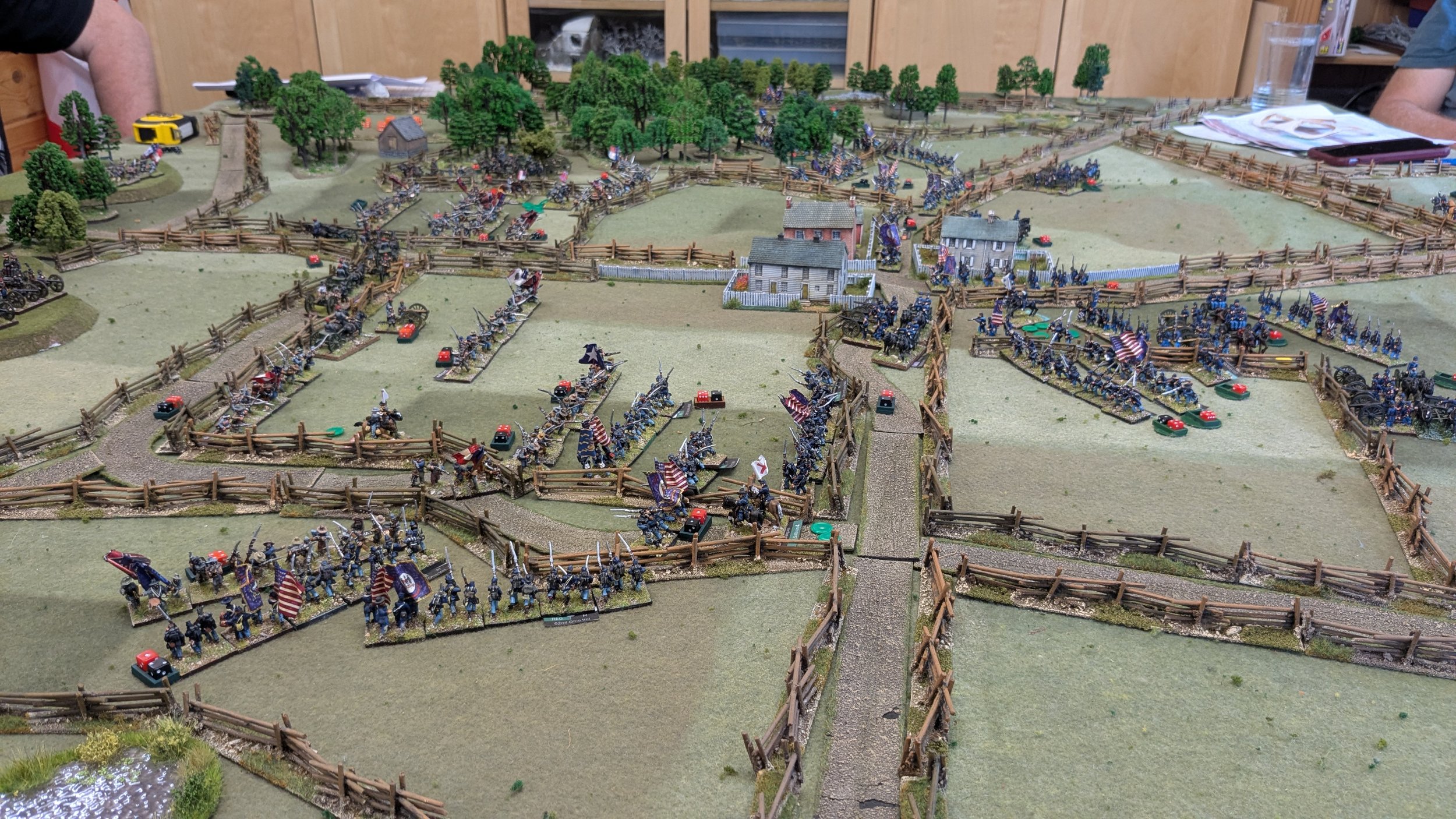 Miniature diorama of a historical battlefield with soldiers fighting behind wooden fences, trees, and small houses, depicting a Civil War or Revolutionary War scene.