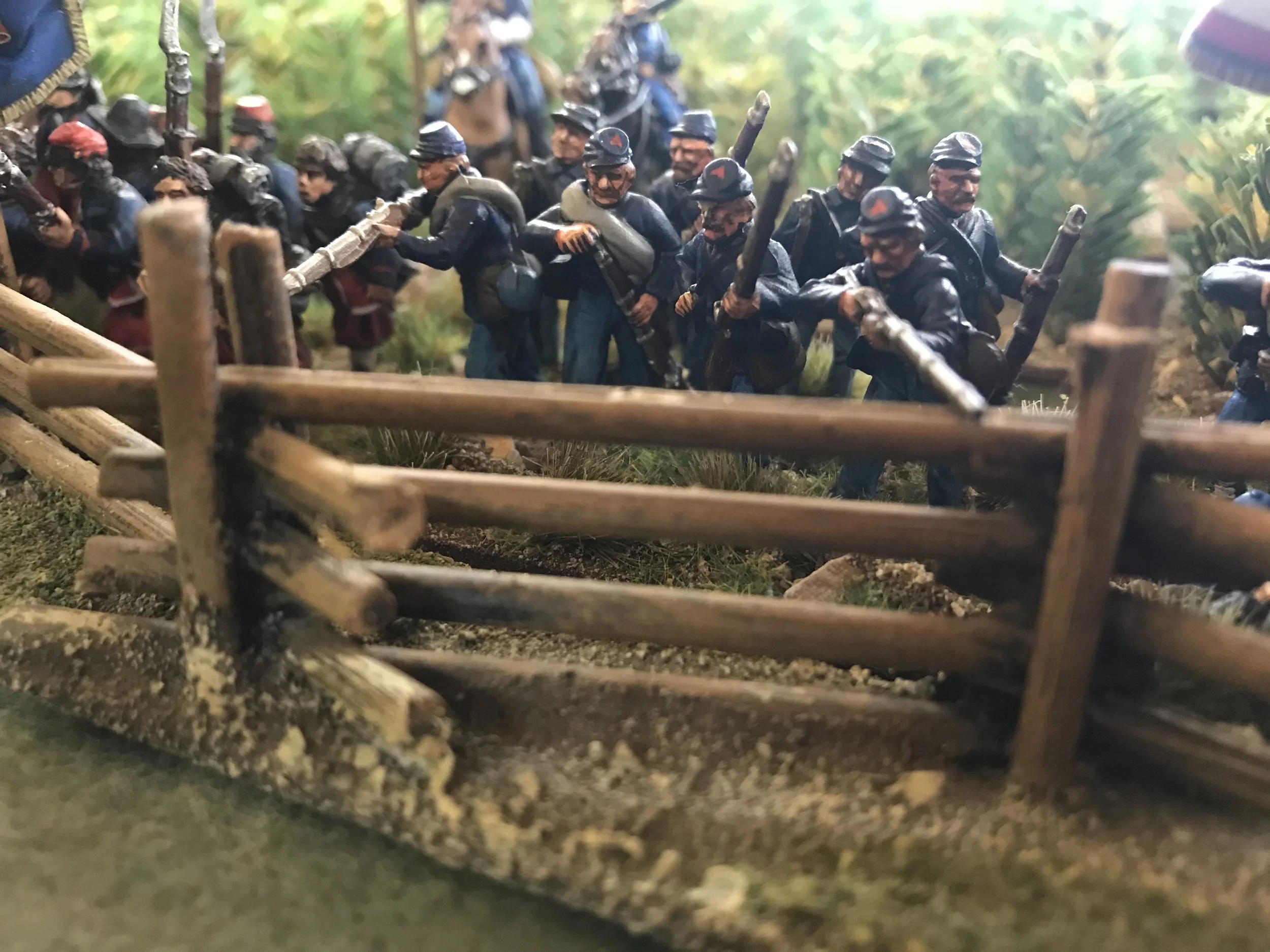Miniature figures of soldiers in a combat scene, crawling behind a wooden barricade with a green forest background.