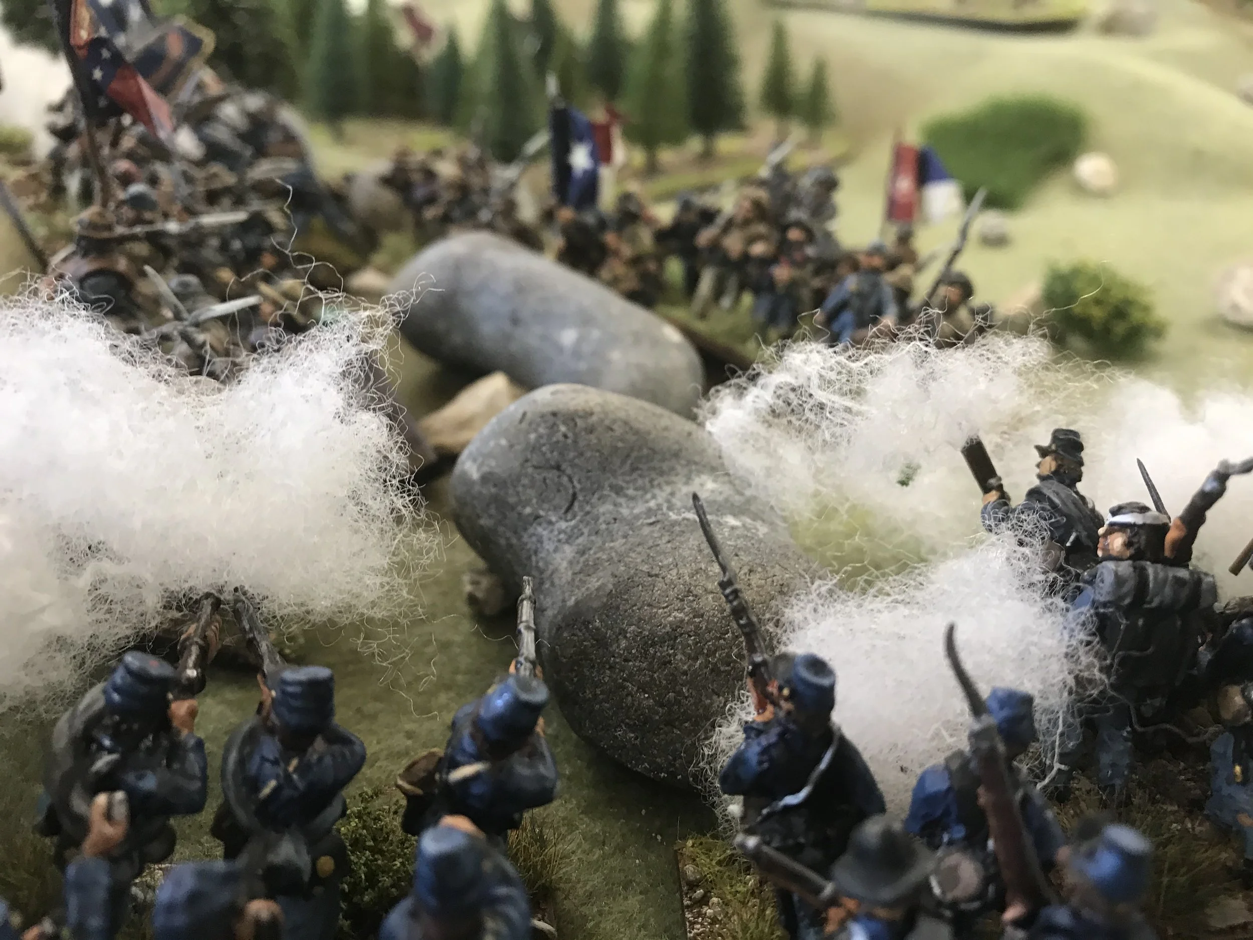 Miniature toy soldiers arranged on a surface simulating a battlefield, with some soldiers hiding behind rocks and bushes, and smoke effects among the figures.