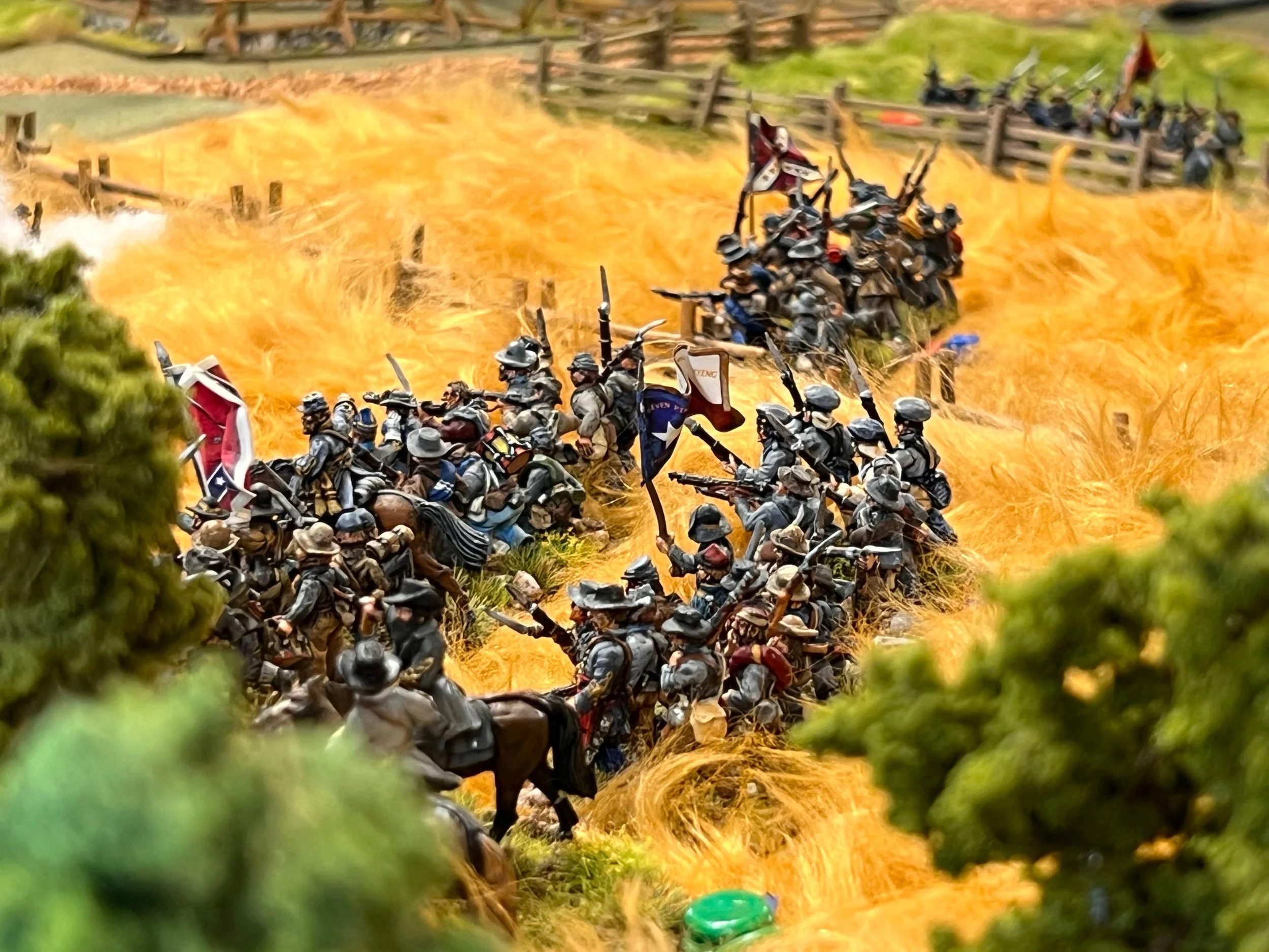 Miniature scene of soldiers in uniform and helmets holding rifles, engaged in a battle with explosions and fire in the background, viewed through green foliage.