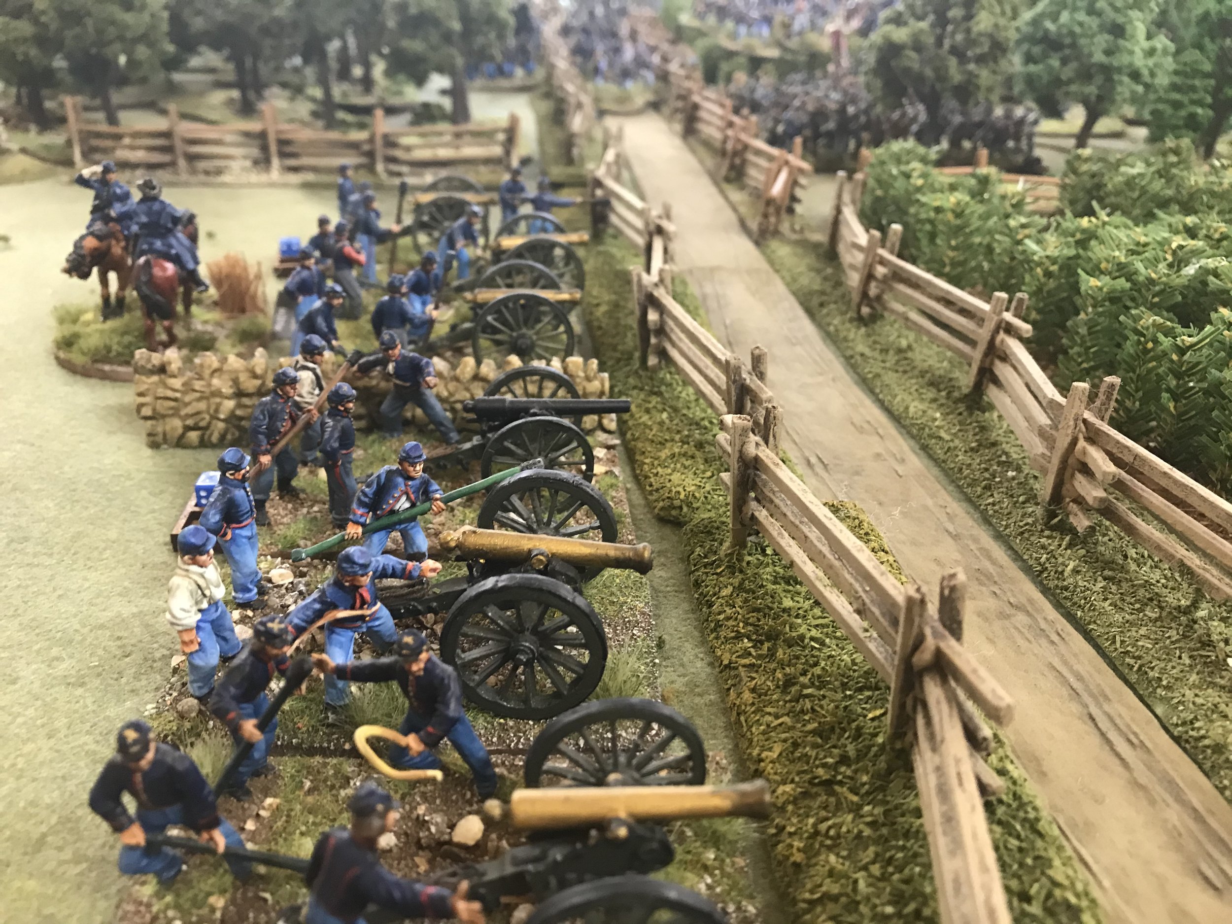 Miniature models of soldiers, in blue uniforms, operating cannons along a dirt path with wooden fences and trees in the background.