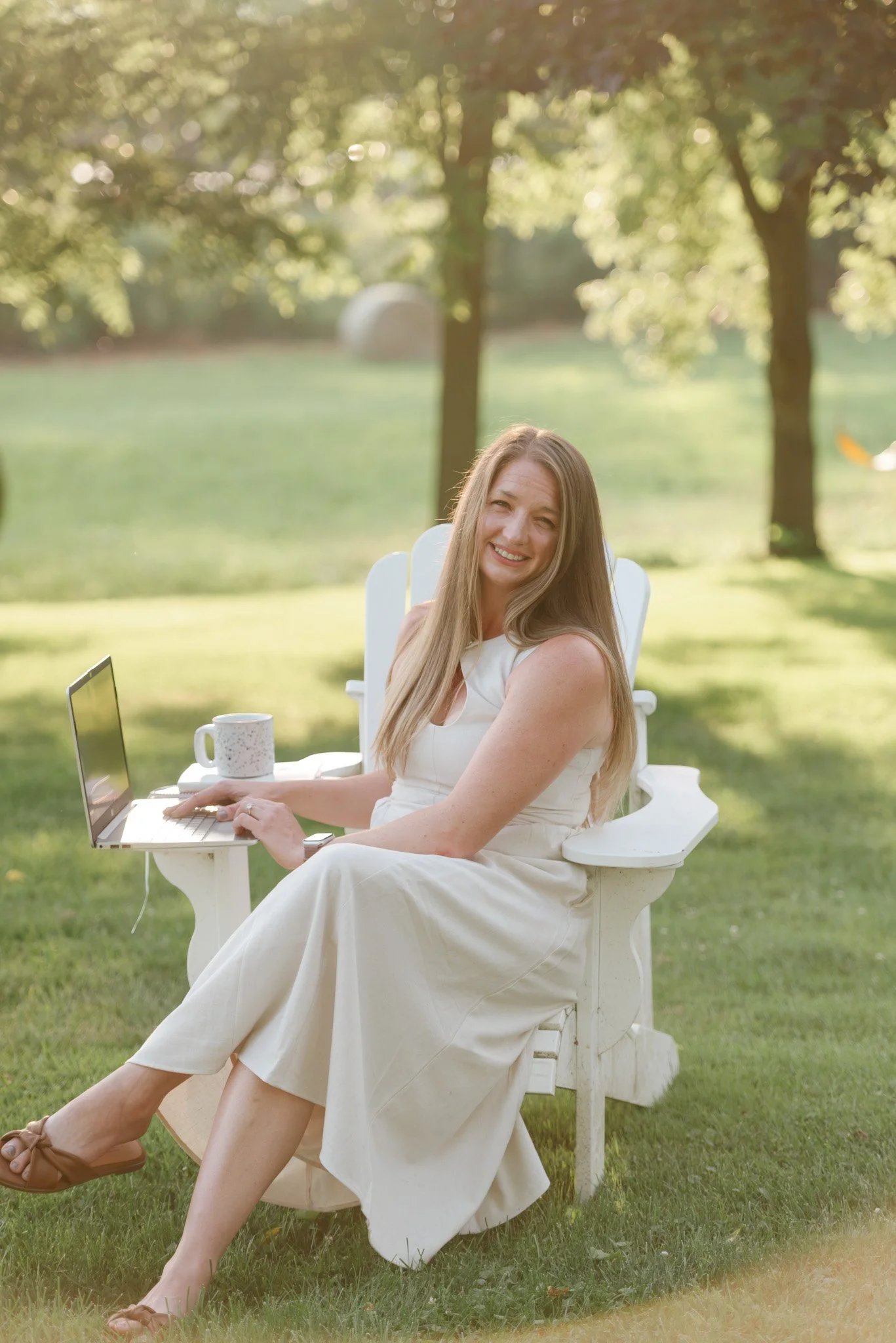 A woman with long blonde hair sitting on a white outdoor chair in a grassy area, smiling, with a laptop on her lap, a mug on a small white table, and trees in the background during daylight.