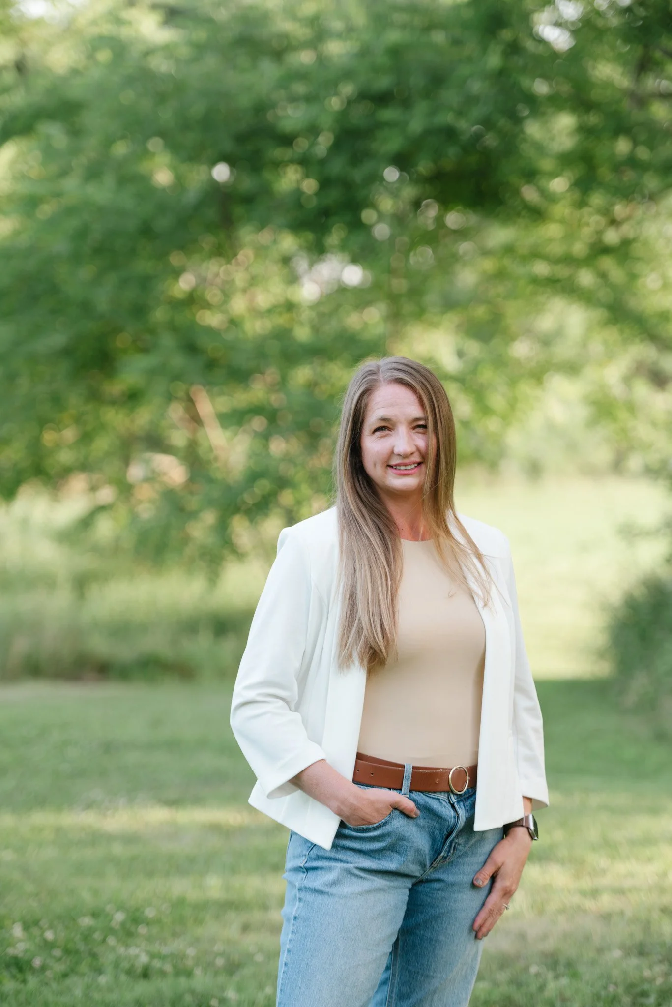 Young woman in beige top and white blazer, with long blonde hair, standing outdoors on a grassy field with green trees in the background.