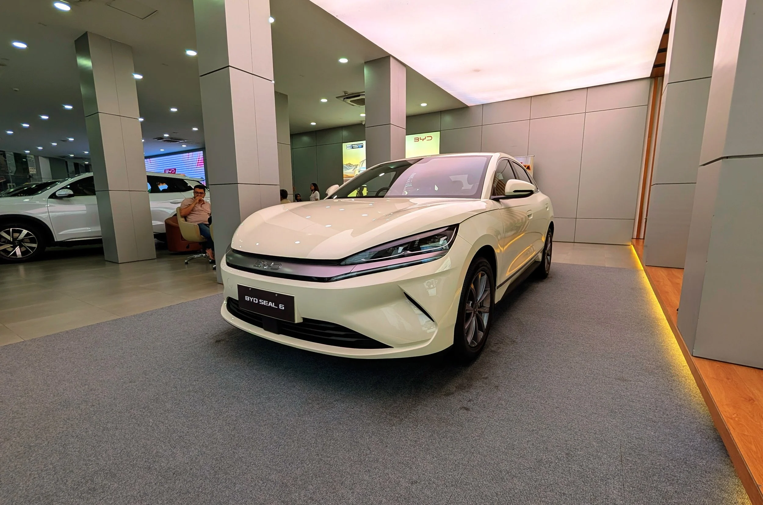 A white BYD Seal 6 electric car is on display in BYD Flagship showroom in Dhaka, with other cars and people in the background.