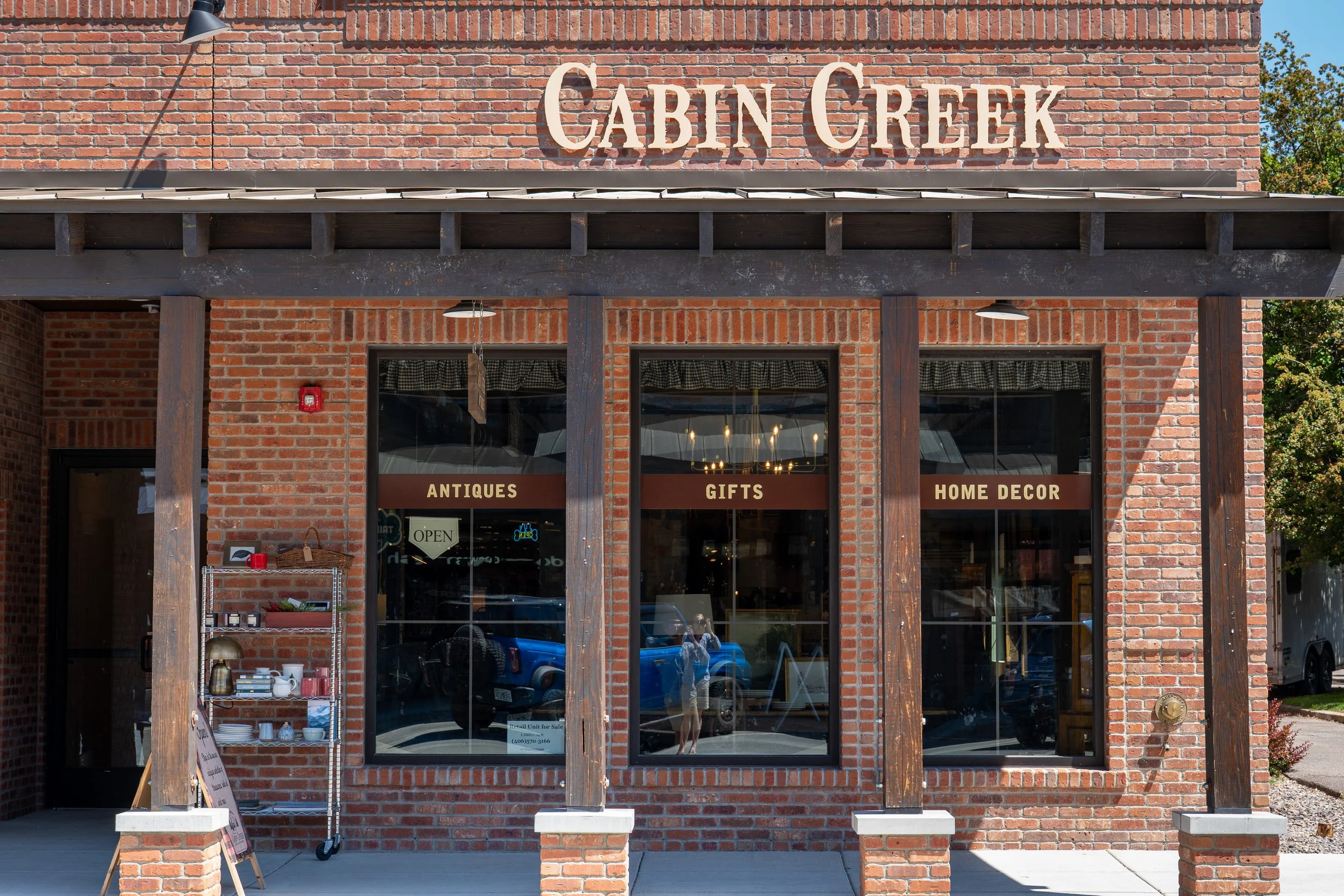 The storefront of a shop named 'Cabin Creek' with a brick exterior, large windows, and dark brown wooden accents. Signs on the windows indicate it sells antiques, gifts, and home decor. There is a small display shelf outside with various items and a sign indicating a retail unit for sale.